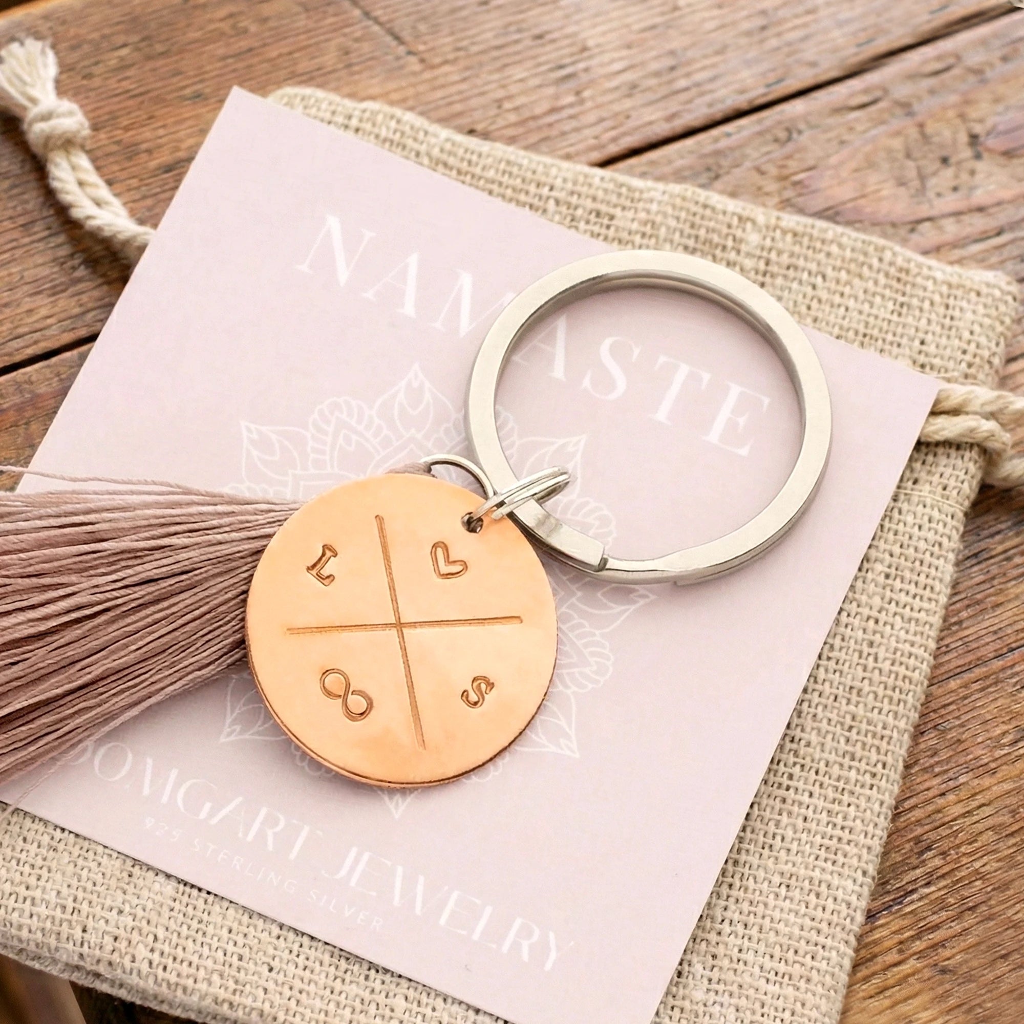 Ein personalisierter Schlüsselanhänger aus Kupfer mit Initialen, Herz und Unendlichkeitszeichen, von Hand gestempelt, verpackt im Boho-Säckchen auf einem alten Holztisch liegend.
