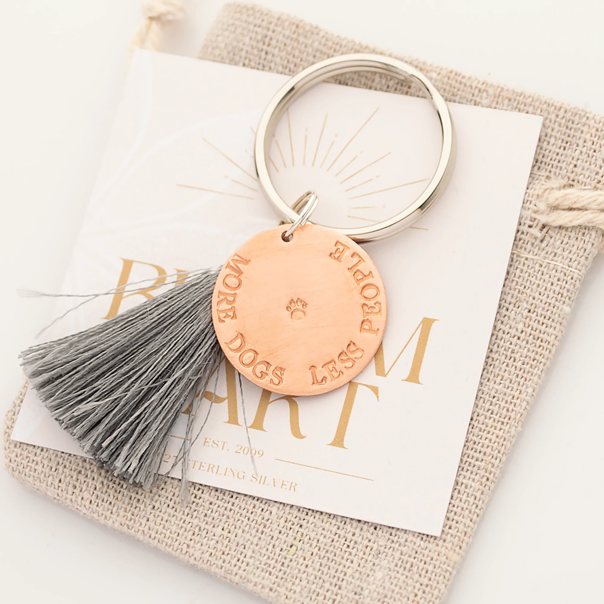 Schlüsselanhänger mit Gravur 'More dogs less people' und einer kleinen Pfote mittig von handgestempelt. Der Schlüsselanhänger liegt verschenk-fertig auf einer Karte und einem beigefarbigen Leinen-Säckchen.
