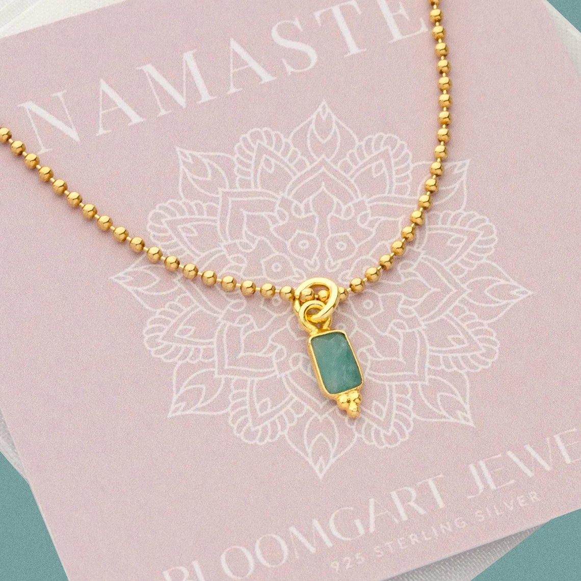 Filigrane goldene Kugelkette mit einem rechteckigen, grün-türkisen Amazonit Anhänger von Bloomgart Jewelry auf einer rosa Produktkarte.