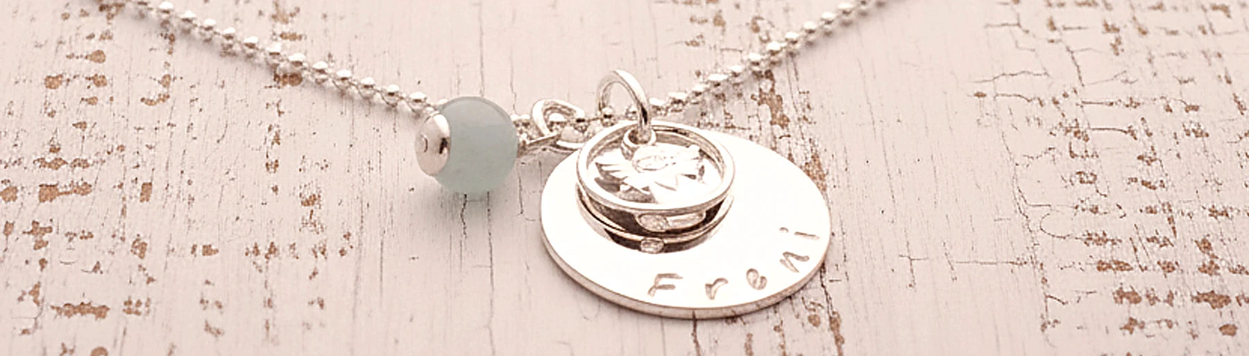Makroansicht einem 925 Silber Taufketterl mit großem Gravurtaler, hier mit dem Namen 'Freni' von Hand gestempelt und einem kleinen Engerl im Taufring. Neben dem Gravurtaler hängt eine Aqua-Chalcedon-Kugel in mint. Die Kette wird auf einem alten Holztisch im shabby-chic Look präsentiert.