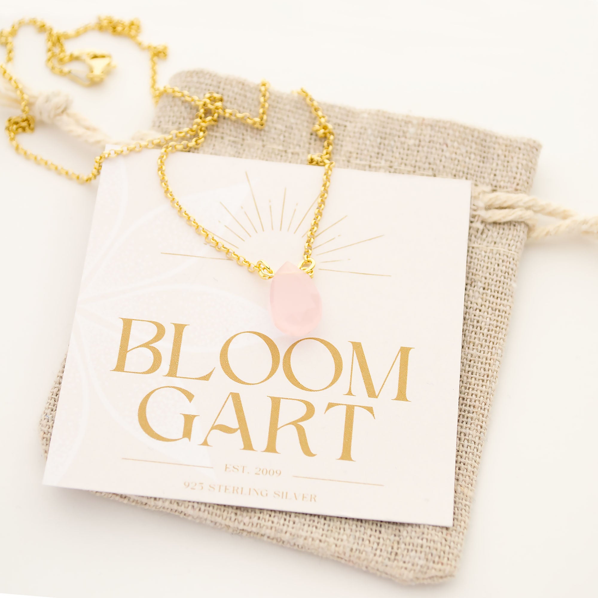 Eine filigrane Kette aus vergoldetem 925 Sterling Silber mit einem Rosenquarz Tropfen-Anhänger. Die Kette liegt auf einer rustikalen Geschenkkarte mit Jute-Säckchen im Boho-Stil.