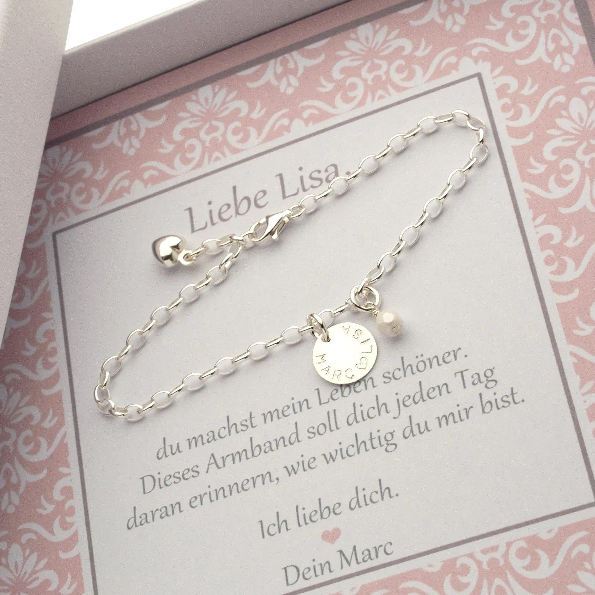 Innenansicht einer Geschenkbox in weiß-rosa zum Jahrestag. Auf einer personalisierten Karte liegt ein 925 Silberarmband mit Gravurtaler "Lisa ♡ Marc" und weißer Perle. Das Armband wird zum Jahrestag an die Freundin verschenkt.