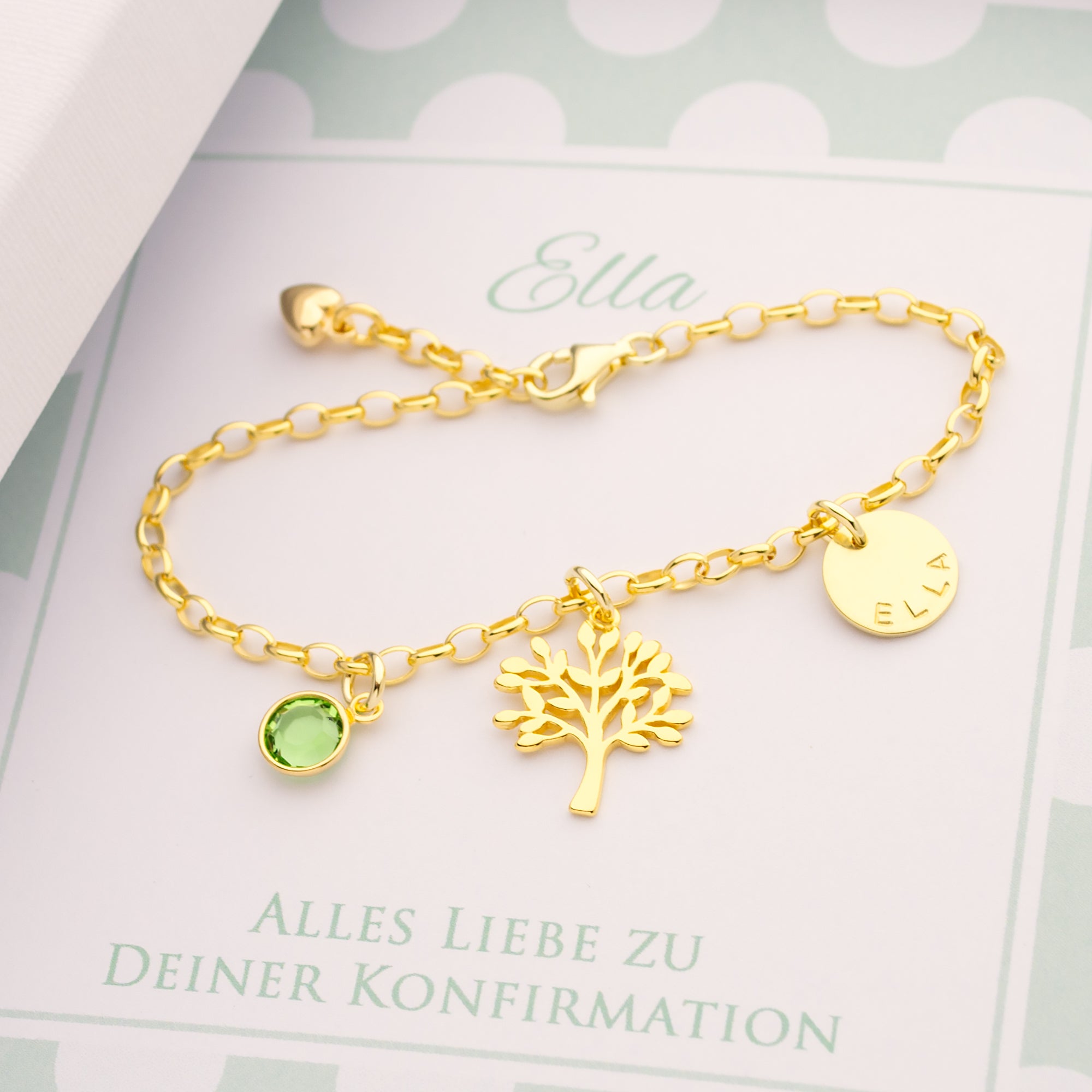 Detailansicht eines personalisierten Namensarmbandes aus 18k vergoldetem 925 Silber, mit Lebensbaum-Anhänger, hellgrünem Geburtsstein und Namenstaler, präsentiert in einer personalisierten mintgrünen Geschenkbox zur Konfirmation von Ella.