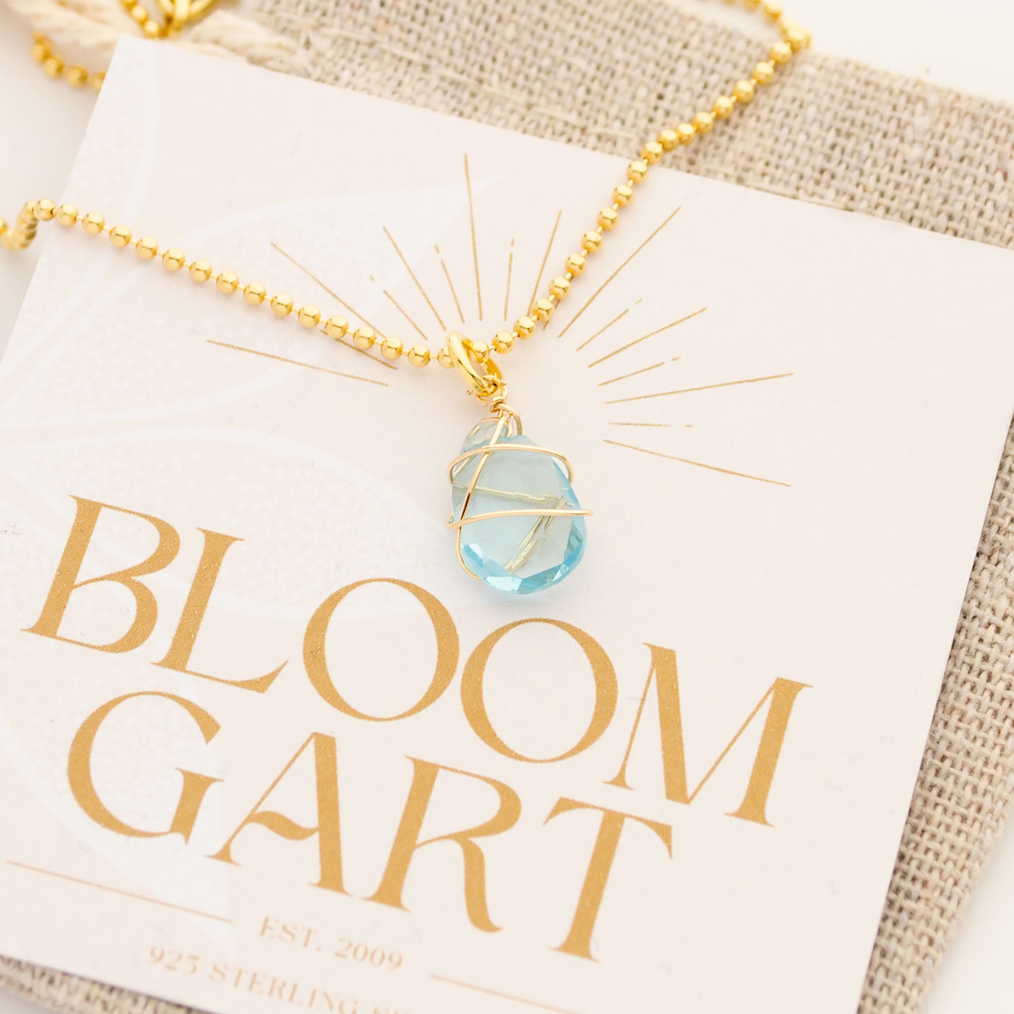 Nahaufnahme einer Goldkette mit einem hellblauen Aqua Chalcedon Tropfen, der kunstvoll von Goldfaden umwickelt ist. Wundervolle Handarbeit im Boho-Stil.