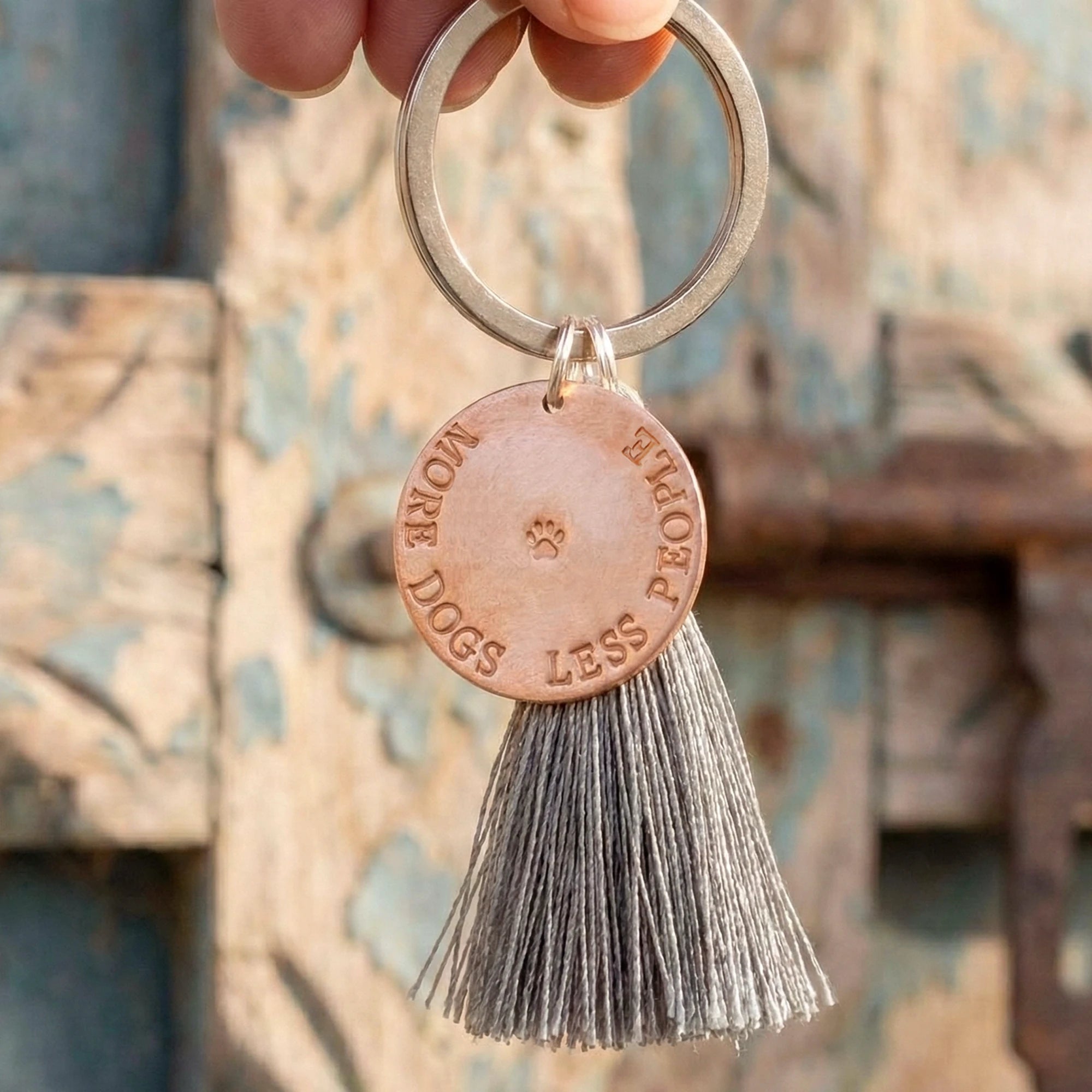 Geschenk für Hundeliebhaber: personalisierter Schlüsselanhänger mit Kupfertaler 'MORE DOGS LESS PEOPLE' und Pfote. Der Schlüsselring baumt an den Fingern einer Hand vor einer alten Holztür.