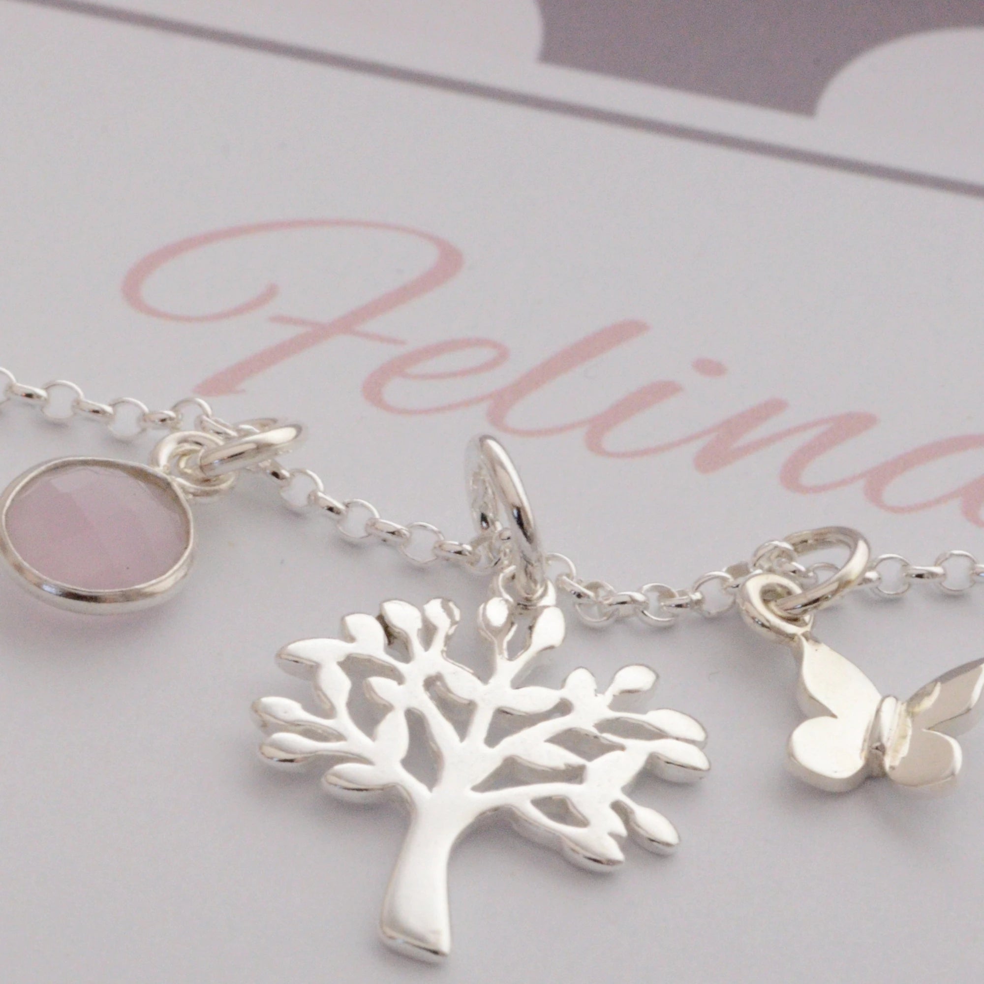 Detailansicht einer filigranen Kette mit drei Anhängern: Rosenquarz in Silber gefasst, Lebensbaum und Schmetterling, aus 925 Sterling Silber. Präsentiert in einer personalisierten Geschenkbox in taupe und rosé.