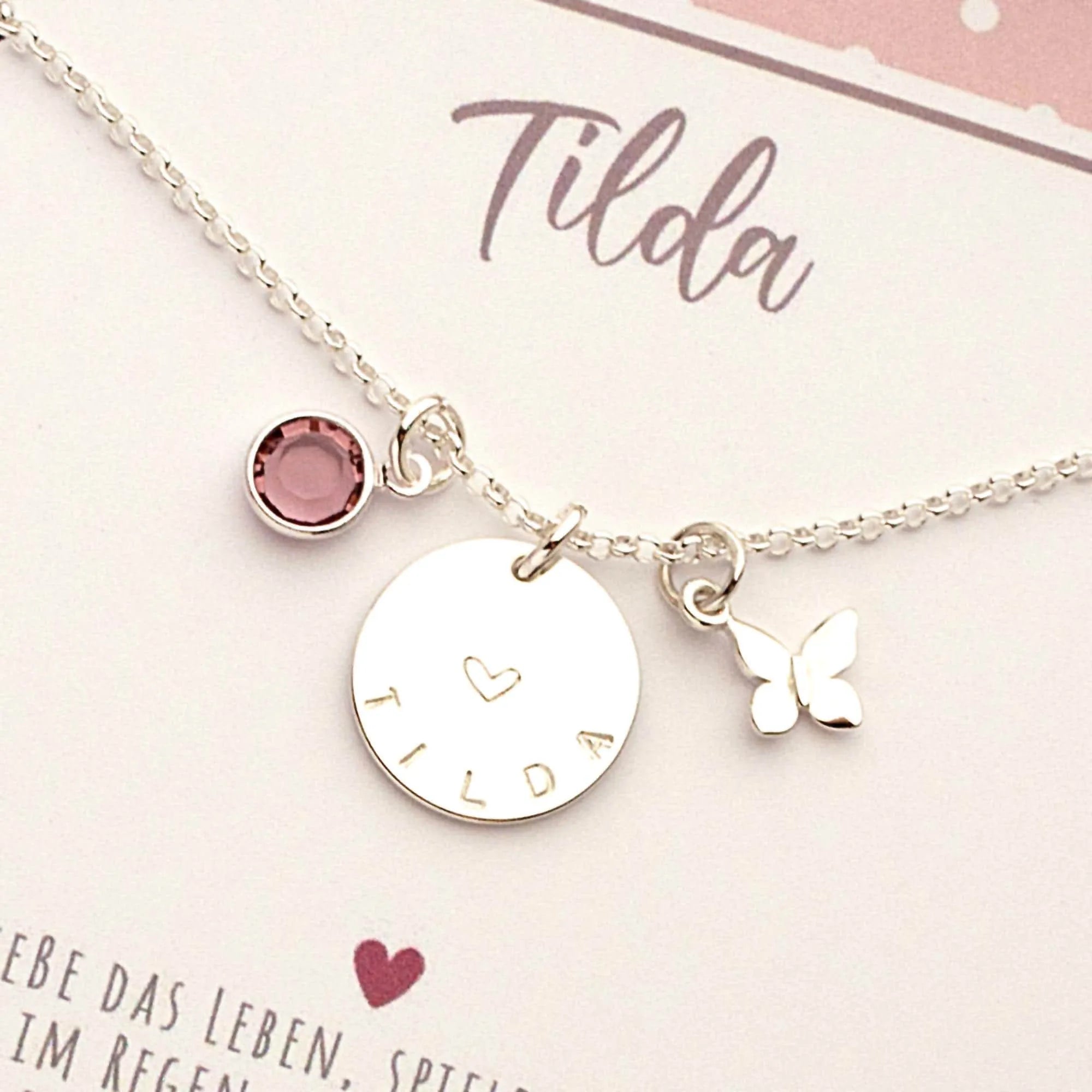 Nahaufnahme einer personalisierten Silberkette für Mädchen mit Gravur 'Tilda', einem Herz-Symbol, Schmetterling-Anhänger und einem flieder-farbigen Geburtsstein. Handgemachtes Geschenk von Bloomgart.