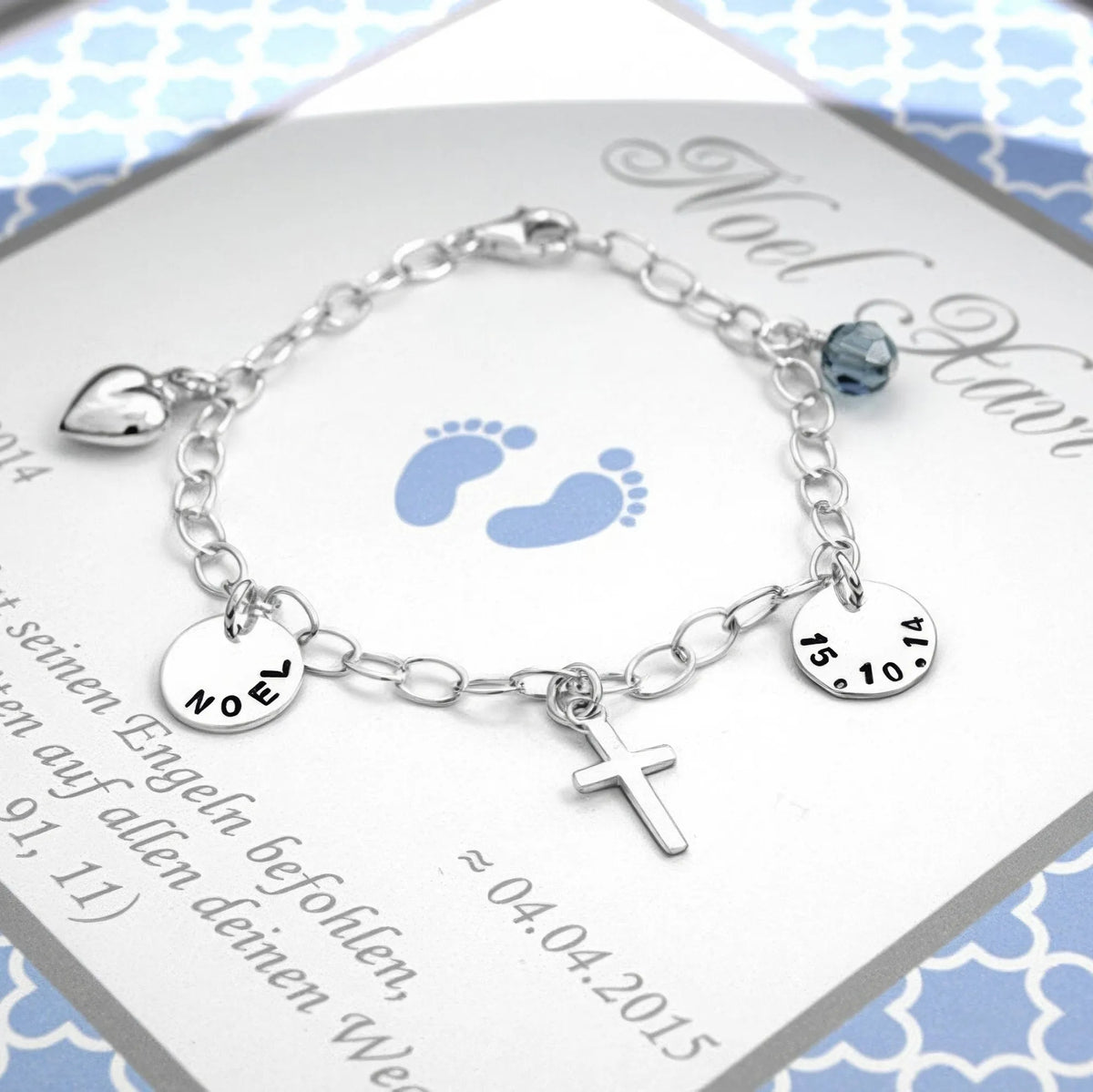 Silberarmband für Jungen mit zwei Gravurtalern für Name und Datum, Herz, Kreuz und Geburtsstein-Kugel auf einer personalisierten Karte in hellblau und weiß.