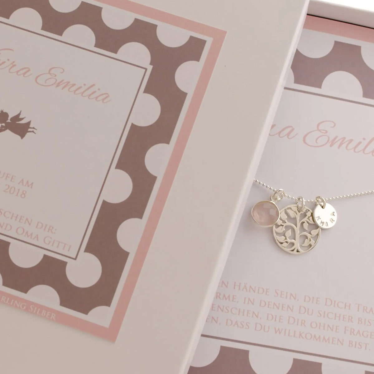 Geschenkbox mit weiß-taupe-rosa farbigem Punkte-Muster, personalisiert für einen besonderen Anlass, hier für 'Khira Emilia' zur Taufe. Innen liegt die handgefertigte Silberkette mit Lebensbaum, Rosenquarz und Namenstaler von Bloomgart.