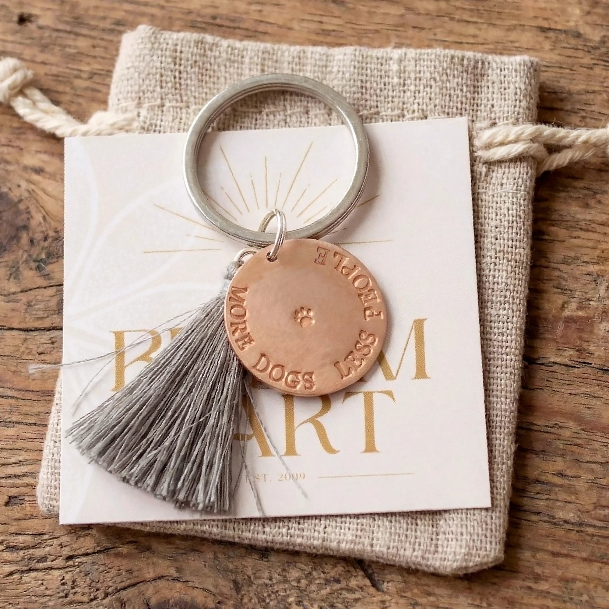 Ein edles Geschenk für Hundebesitzer ist dieser Schlüsselanhänger aus Kupfertaler 'MORE DOGS LESS PEOPLE', einer kleinen Pfote von Hand gestempelt sowie einer grauer Quaste und einem Leinen-Säckchen von Bloomgart. Der Anhänger liegt auf einem alten braunen Holztisch.