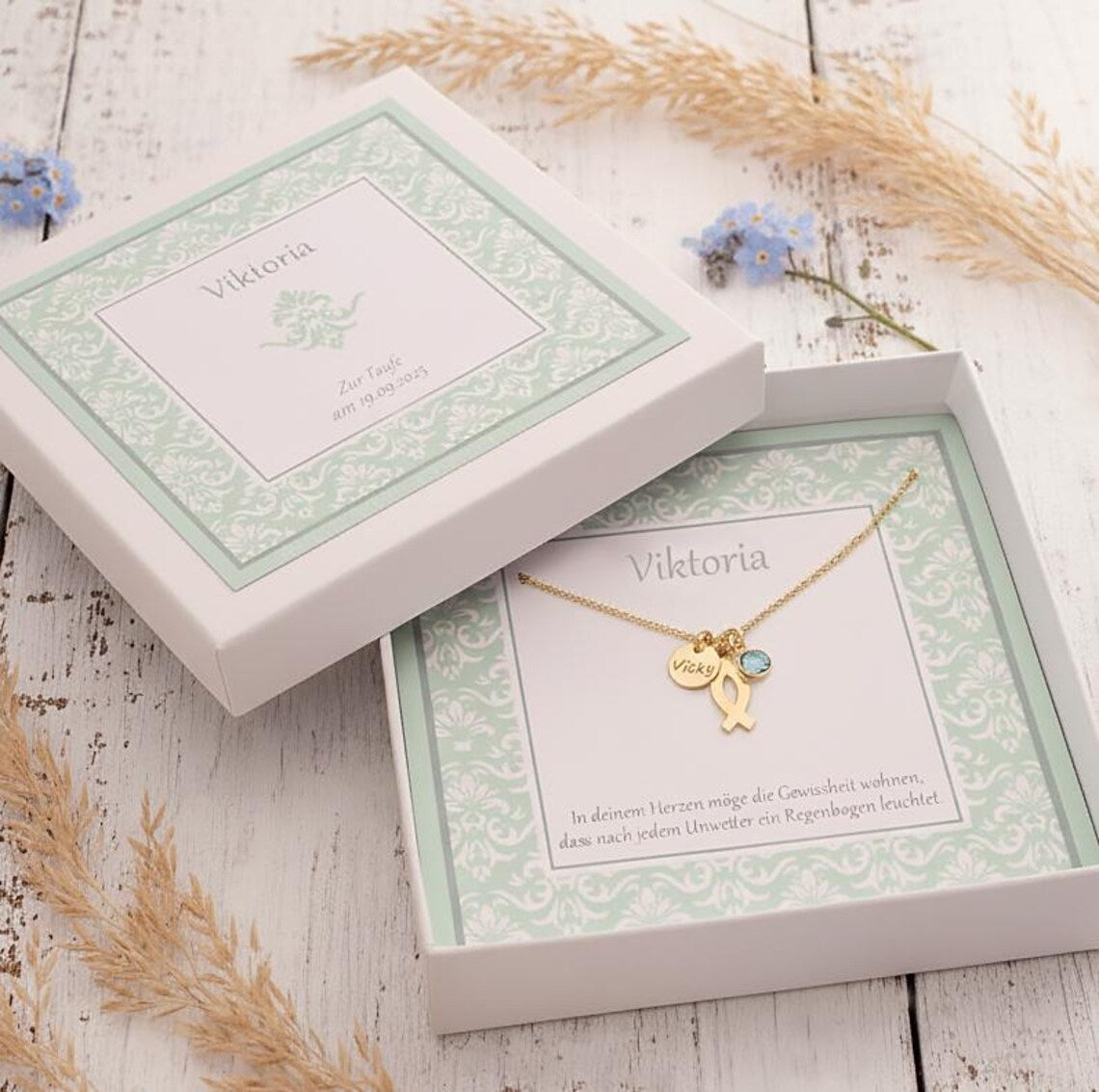 Personalisierte mintgrüne Geschenkbox, in der eine zierliche Goldkette zur Taufe mit Ichthys-Fisch, Monatsstein und Namenstaler 'Vicky' liegt. Die Box ist mit Namen, Anlass und Taufspruch personalisiert und liegt auf einem alten, weißen Vintage-Holztisch mit hellblauen Vergissmeinnicht-Blümchen und Gräsern verziert.