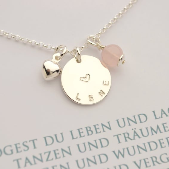 Nahansicht einer zierlichen Silberkette mit bauchigem Herz, großem Namensanhänger, hier 'Lene' sowie einer Rosenquarz-Kugel. Am Verschluss ist ein Datum-Plättchen befestigt.