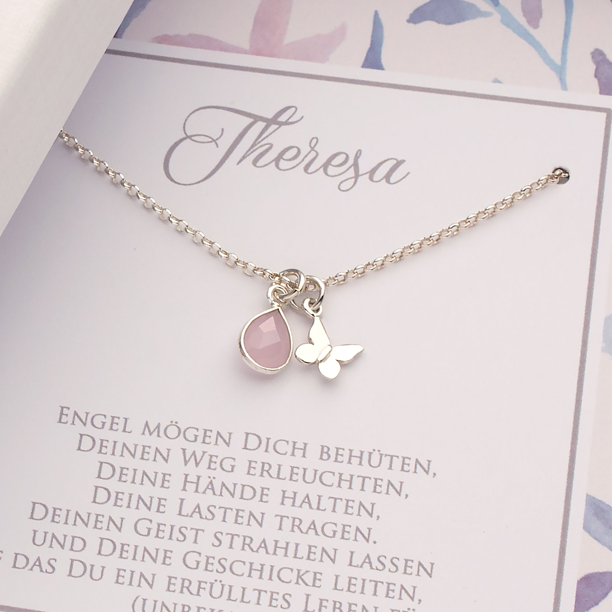 Detailansicht einer filigranen Gliederkette aus 925 Sterling Silber mit Schmetterling und in Silber gefastem Rosenquarz-Tropfen. Oberhalb der Kette ist der Name des Kindes 'Theresa' gedruckt. Unterhalb der Kette ist Platz für einen persönlichen Taufspruch.