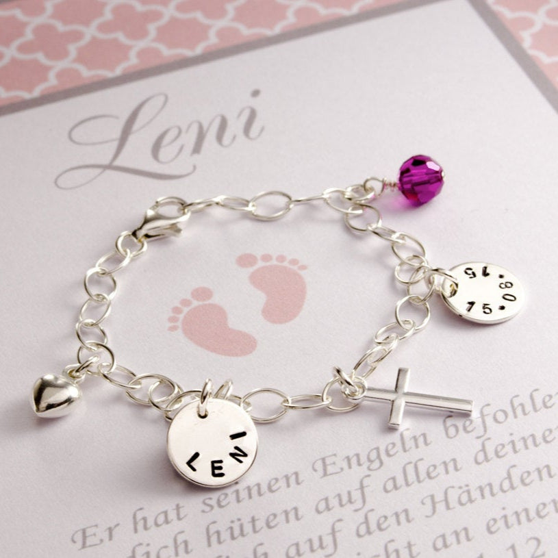Detailansicht eines Silberarmbandes mit Namens- und Datumgravurtaler, Herz, Kreuz und buntem Stein-Anhänger. Das Gliederarmband wird in einer personalisierten Geschenkbox in rosa-weiß präsentiert.