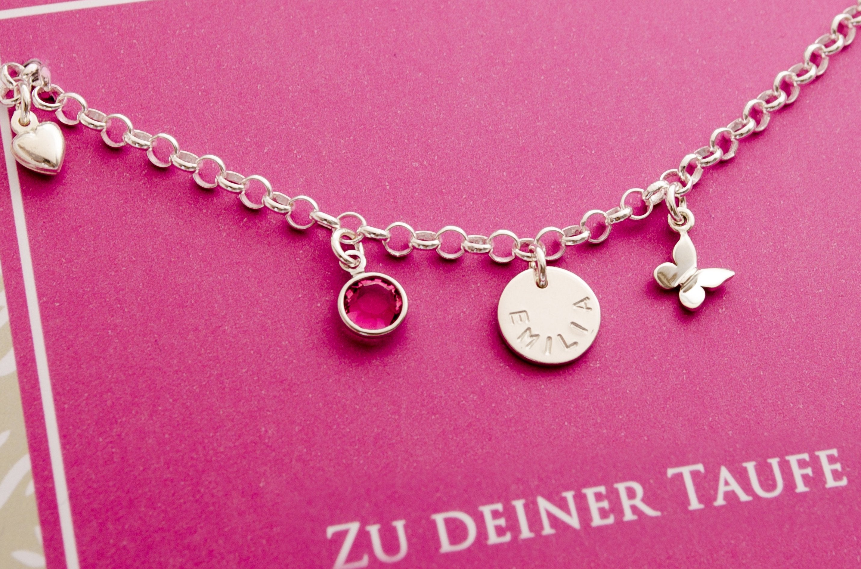 Detailansicht eines Silberarmbandes mit weiten verstellbaren Gliedern zum Mitwachsen mit Namenstaler, Schmetterling und Monatsstein auf einer pink farbigen Unterlage