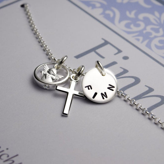 Detailansicht einer Silberkette mit Namensanhänger, Kreuz und Schutzengel im Taufring. Das Taufgeschenk für Jungen wird in einer hellblauen, personalisierten Geschenkbox präsentiert.