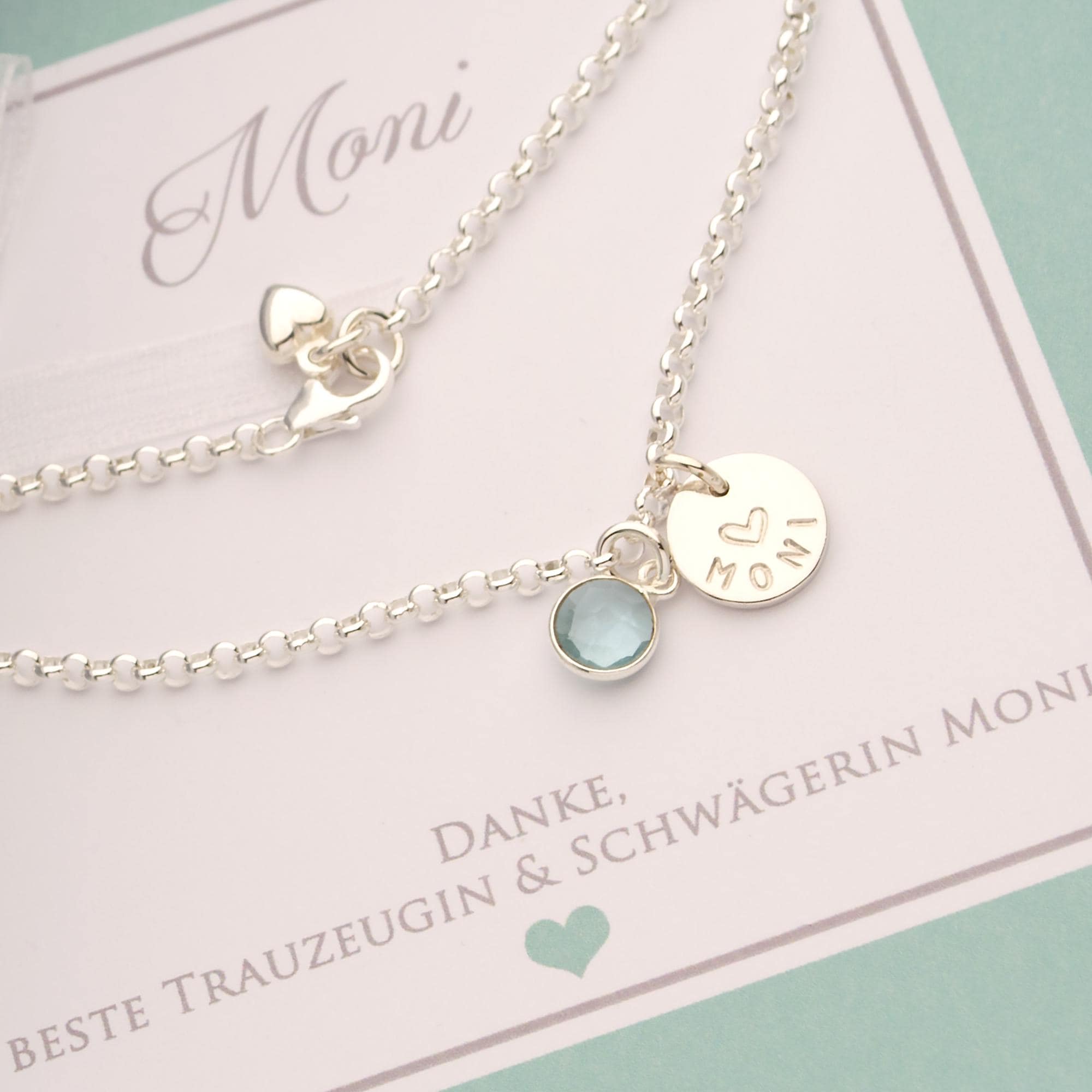 Nahaufnahme eines personalisierten 925 Silberarmbands als Geschenk für die Trauzeugin. Das Plättchen ist mit dem Namen 'Moni' und einem Herz graviert, daneben ein blauer Edelstein. Von Bloomgart.