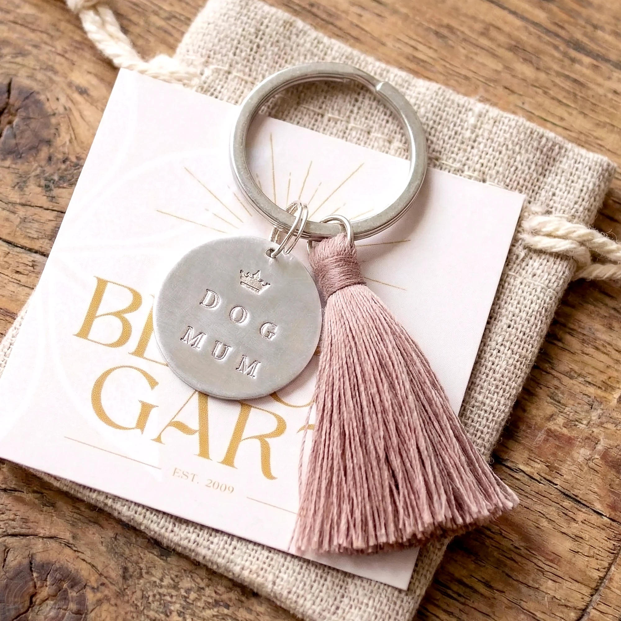 Ein edles Geschenk für Hundebesitzer ist dieser Schlüsselanhänger aus Gravurtaler mit Gravur 'DOG MUM' und einer kleinen Krone sowie einem Quasten-Anhänger aus Kunstseide. Der Anhänger liegt auf einem alten braunen Holztisch.