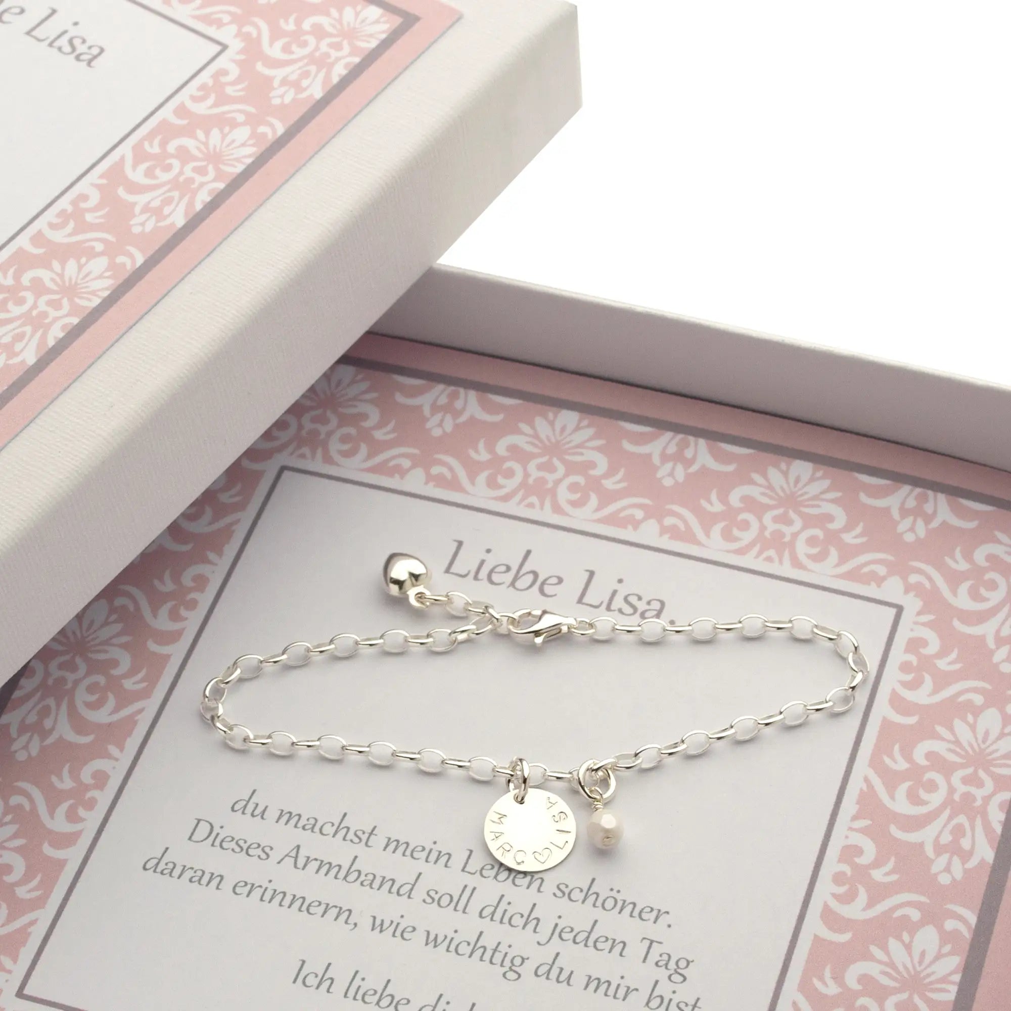 Innenansicht einer weiße Geschenkbox mit rosafarbenem Ornamentmuster und einem 925 Silber-Armband mit Gravur "Lisa ♡ Marc" und weißer Perle. Die Box enthält eine Karte mit der Aufschrift "Liebe Lisa" und "du machst mein Leben schöner. ... Ich liebe dich. Marc".
