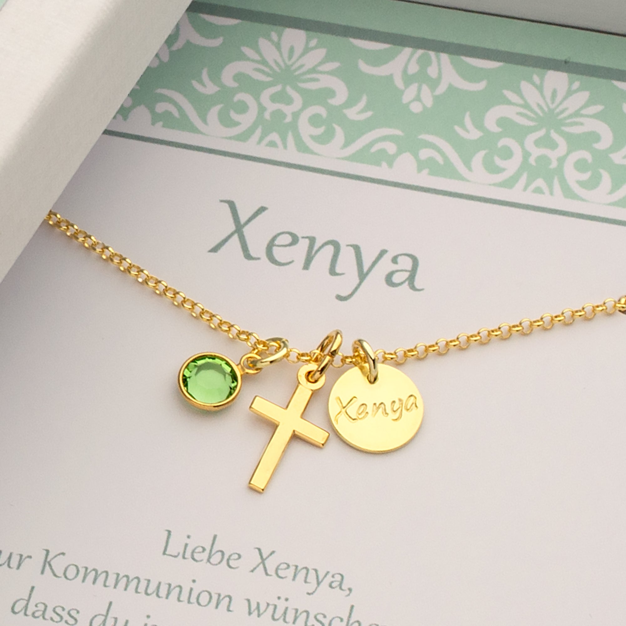 Eine goldene Kette mit den Anhängern Kreuz, Geburtsstein in Wunschfarbe und Namenstaler in einer personalisierten Geschenkbox mit Name, Datum, Anlass und Taufspruch oder Wünschen der Schenkenden.