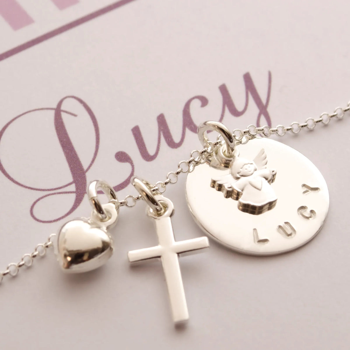 Detailansicht einer filigranen Namenskette mit Schutzengel, Kreuz und Herz-Anhänger, personalisiert für 'Lucy'.
