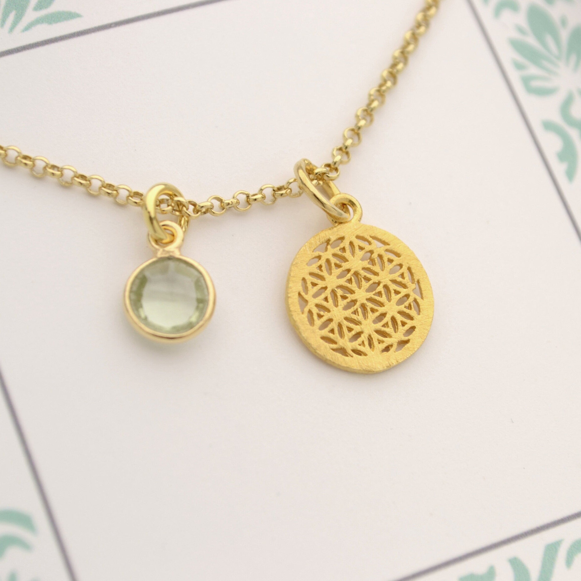 Nahaufnahme einer vergoldete Kette mit Lebensblume-Anhänger und einem grünen Amethyst-Edelstein, präsentiert in einer kleinen Geschenkbox mit Ornament-Muster in mint und taupe.