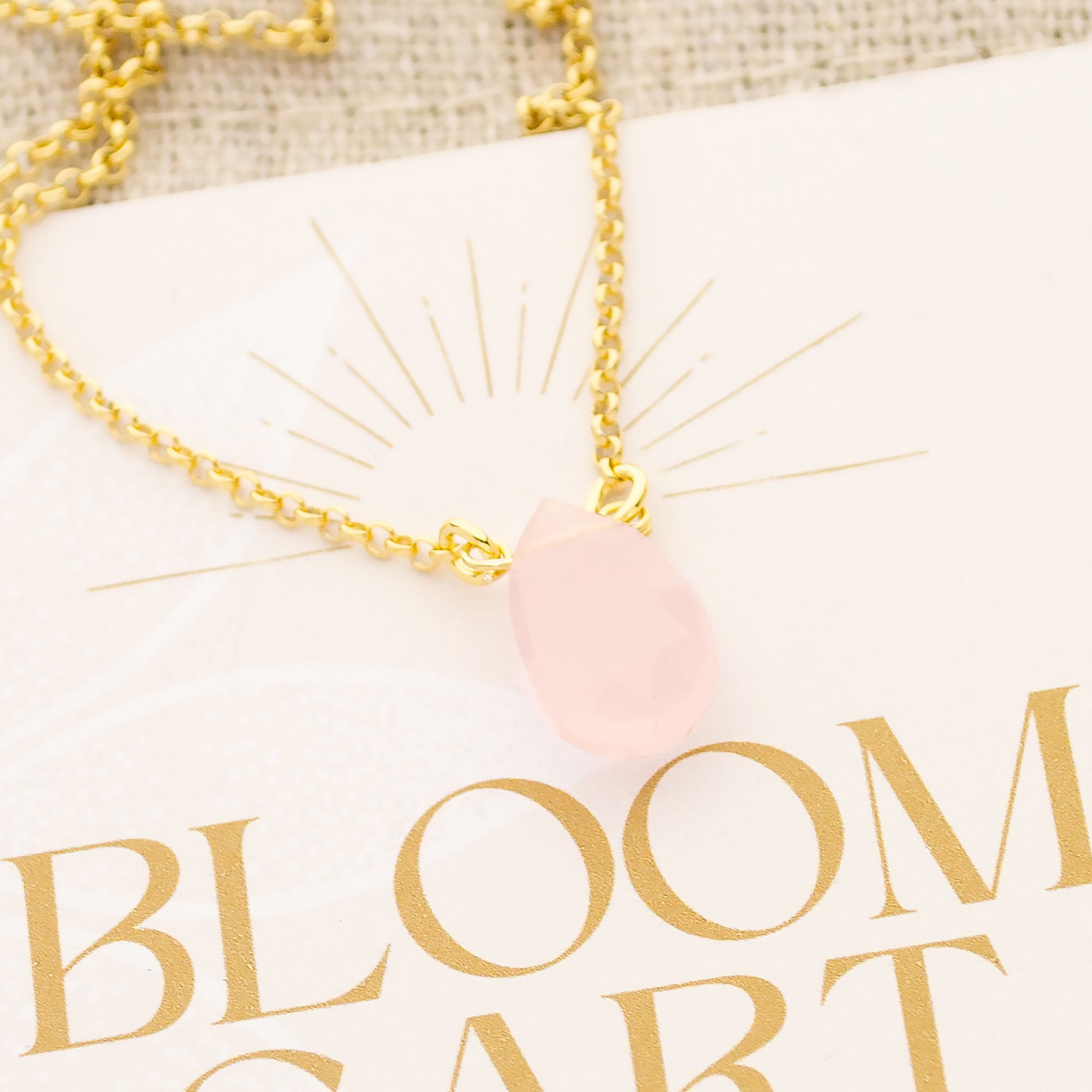 Nahaufnahme einer edlen Goldkette mit einem zart rosa Rosenquarz Tropfen-Anhänger, der mit Draht an der Kette befestigt ist. Die Kette liegt auf einer rustikalen Geschenkkarte mit Jute-Säckchen im Boho-Stil.