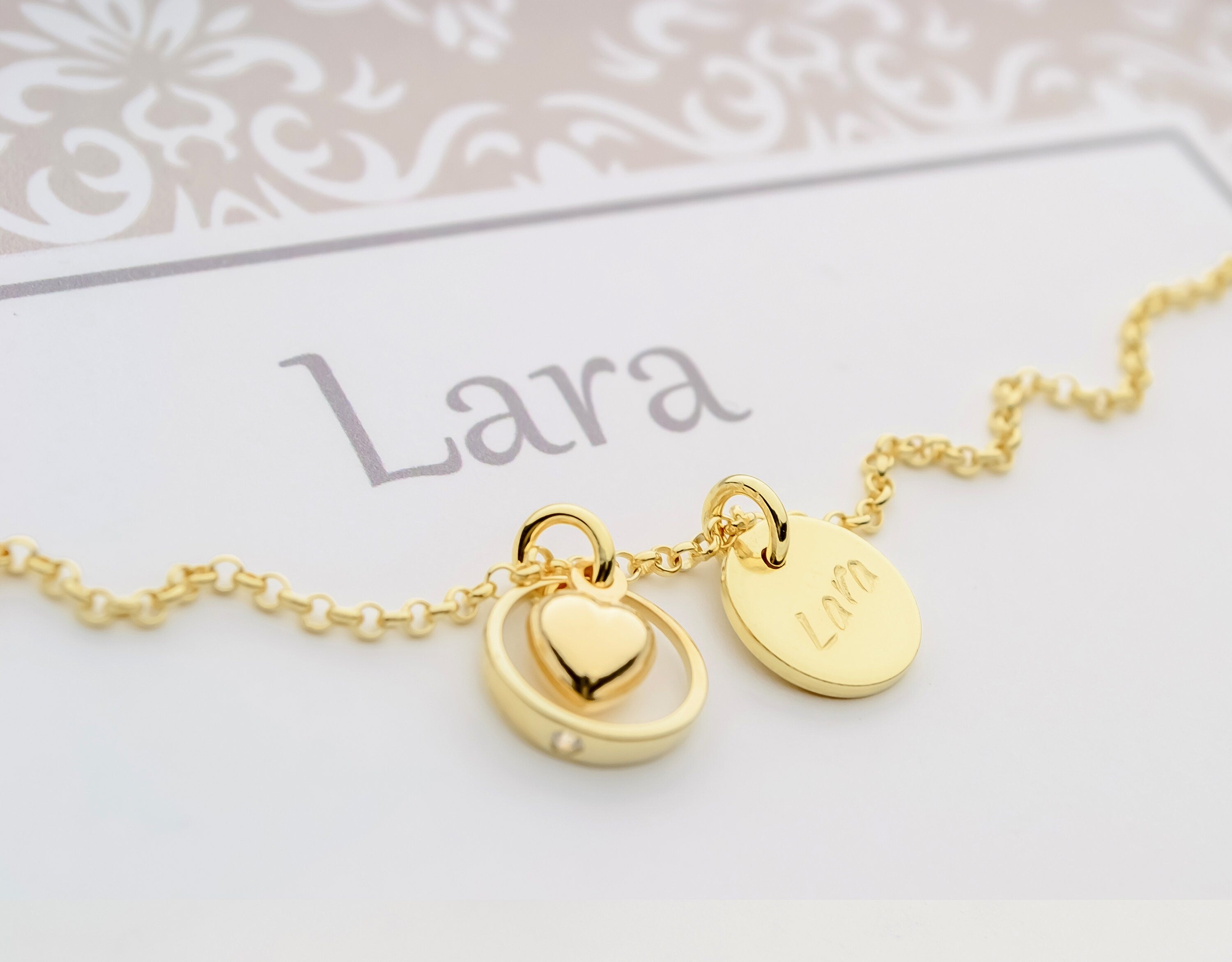 Detailansicht einer goldenen Gliederkette mit zwei Anhängern: einem runden Namenstaler 'Lara' und einem bauchigen Herz im Taufring.