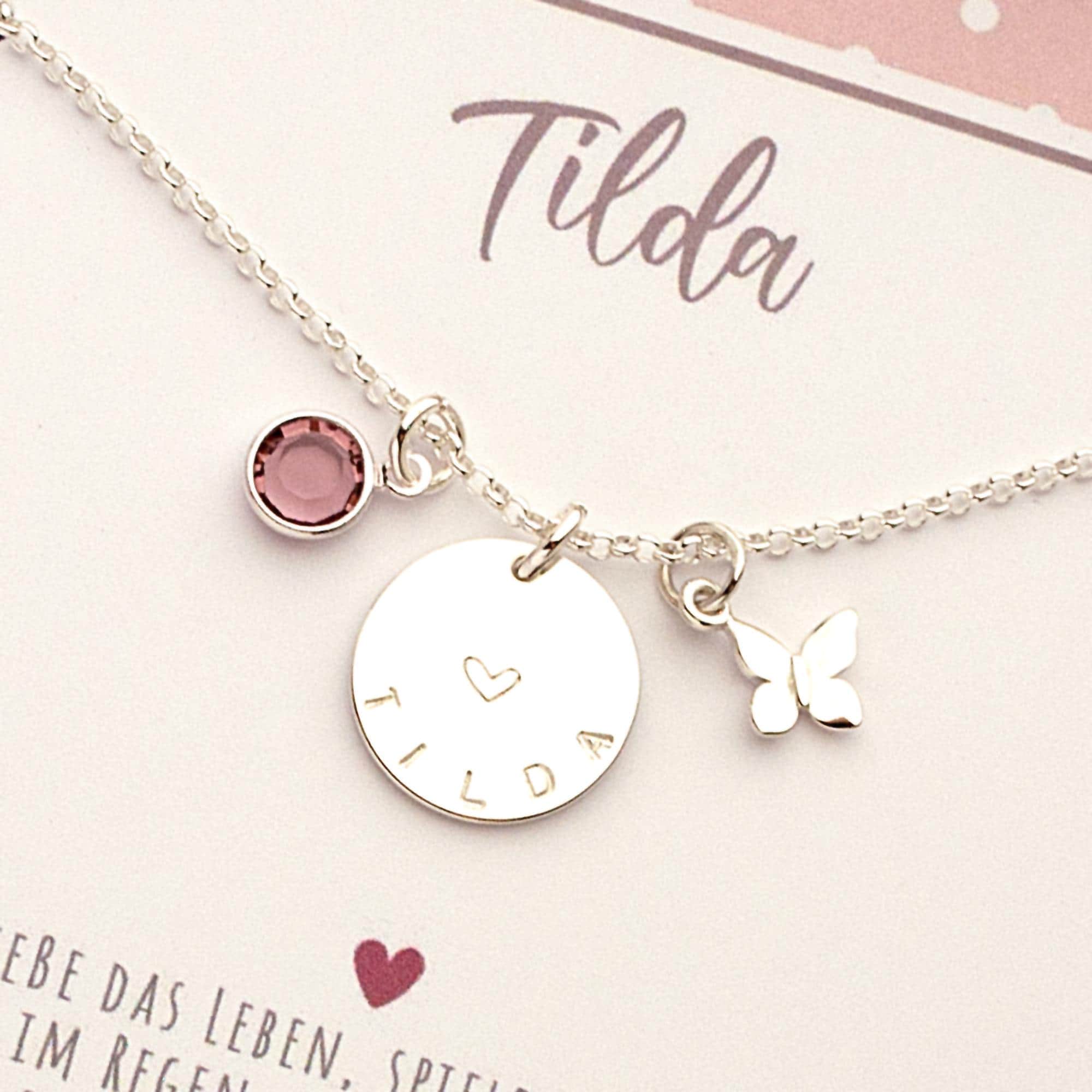 Nahaufnahme einer personalisierten Silberkette für Mädchen mit Gravur 'Tilda', einem Herz-Symbol, Schmetterling-Anhänger und einem flieder-farbenem Geburtsstein. Handgemachtes Geschenk von Bloomgart.