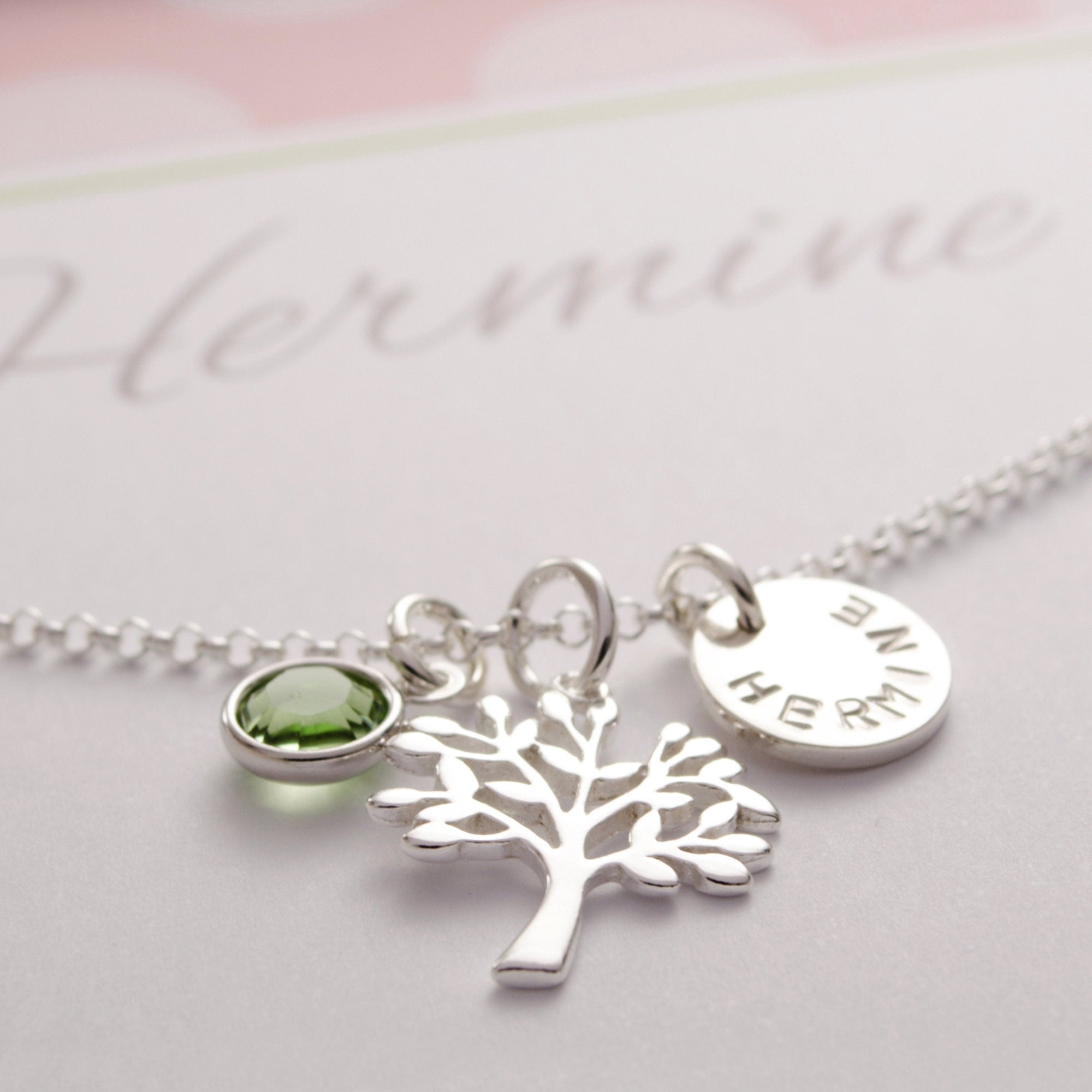 Nahaufnahme einer Lebensbaumkette aus 925 Sterlingsilber. An der Halskette hängen drei Anhänger: ein peridotgrüner August Stein, ein filigraner Lebensbaum und ein Namensplättchen mit 10 mm Durchmesser mit dem Namen "HERMINE".