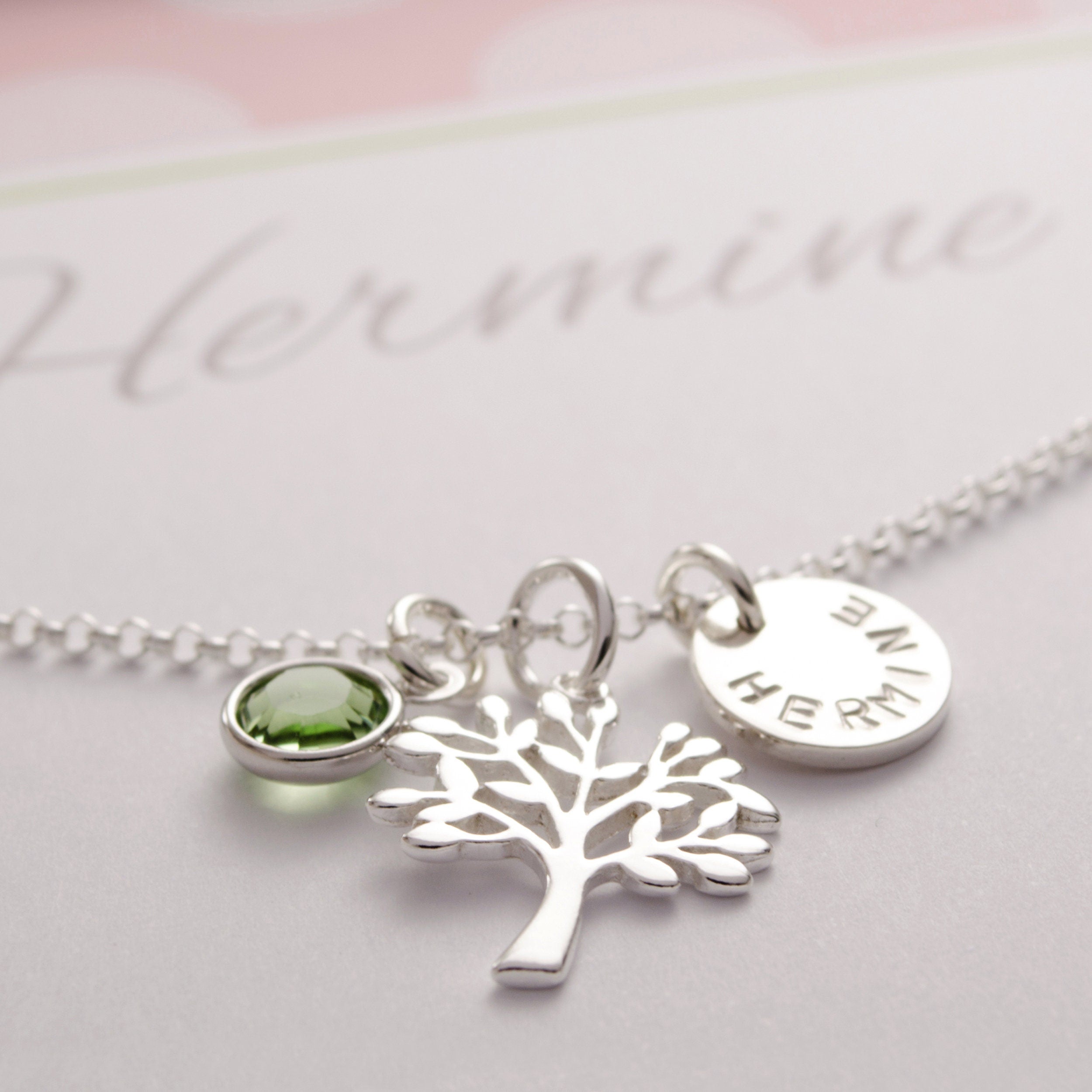 Nahaufnahme einer Lebensbaumkette aus 925 Sterlingsilber. An der Halskette hängen drei Anhänger: ein peridotgrüner August Stein, ein filigraner Lebensbaum und ein Namensplättchen mit 10 mm Durchmesser mit dem Namen "HERMINE".