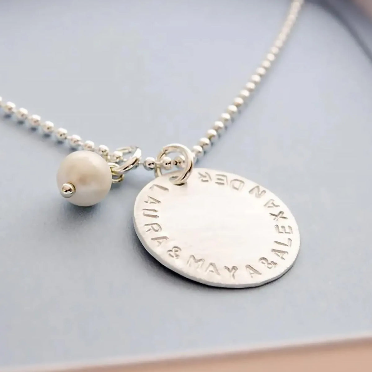 Detailansicht einer Kugelkette mit 18 mm Gravur-Anhänger mit von Hand gestempelten Namen "LAURA & MAYA & ALEXANDER" in einer kleinen hellblauen Geschenkbox