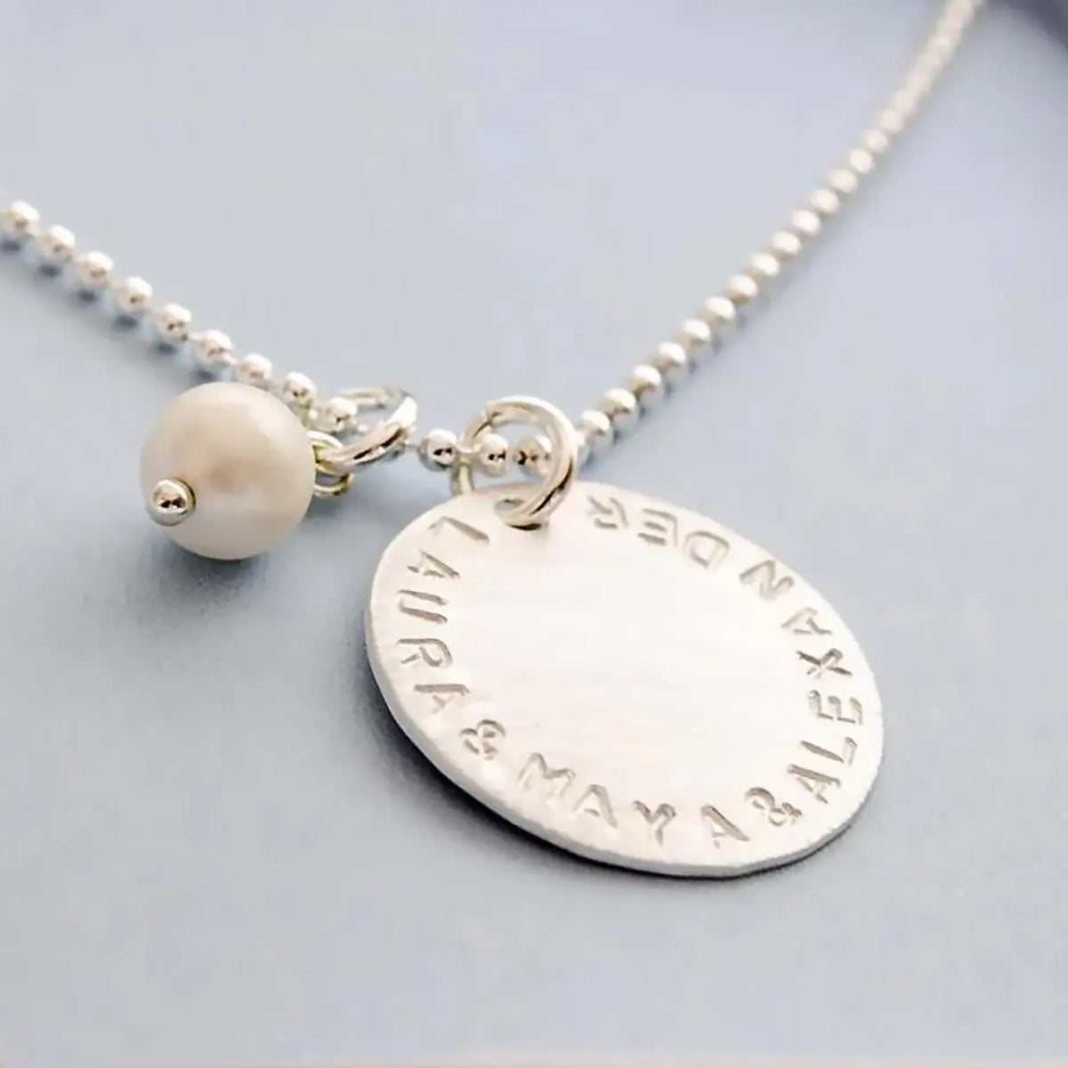 Detailansicht einer 925 Sterling Silber Halskette mit 18 mm Gravur-Anhänger und handgestempelten Namen "LAURA & MAYA & ALEXANDER" in einer kleinen hellblauen Geschenkbox.