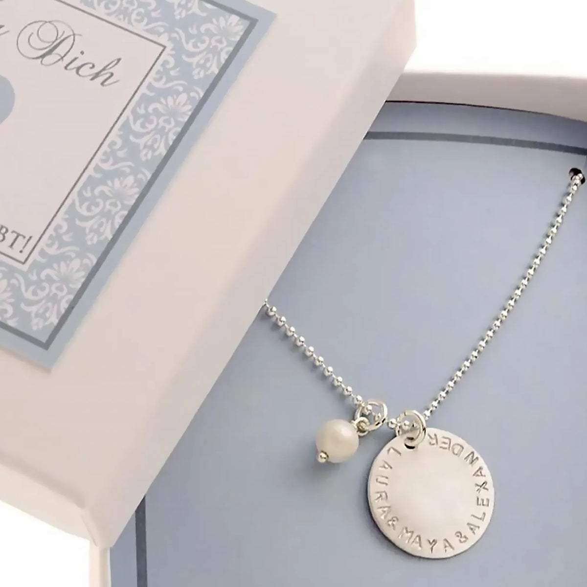 Kugelkette mit 18 mm Wunschgravur Taler mit von Hand gestempelten Namen "LAURA & MAYA & ALEXANDER" in einer kleinen hellblauen Geschenkbox.