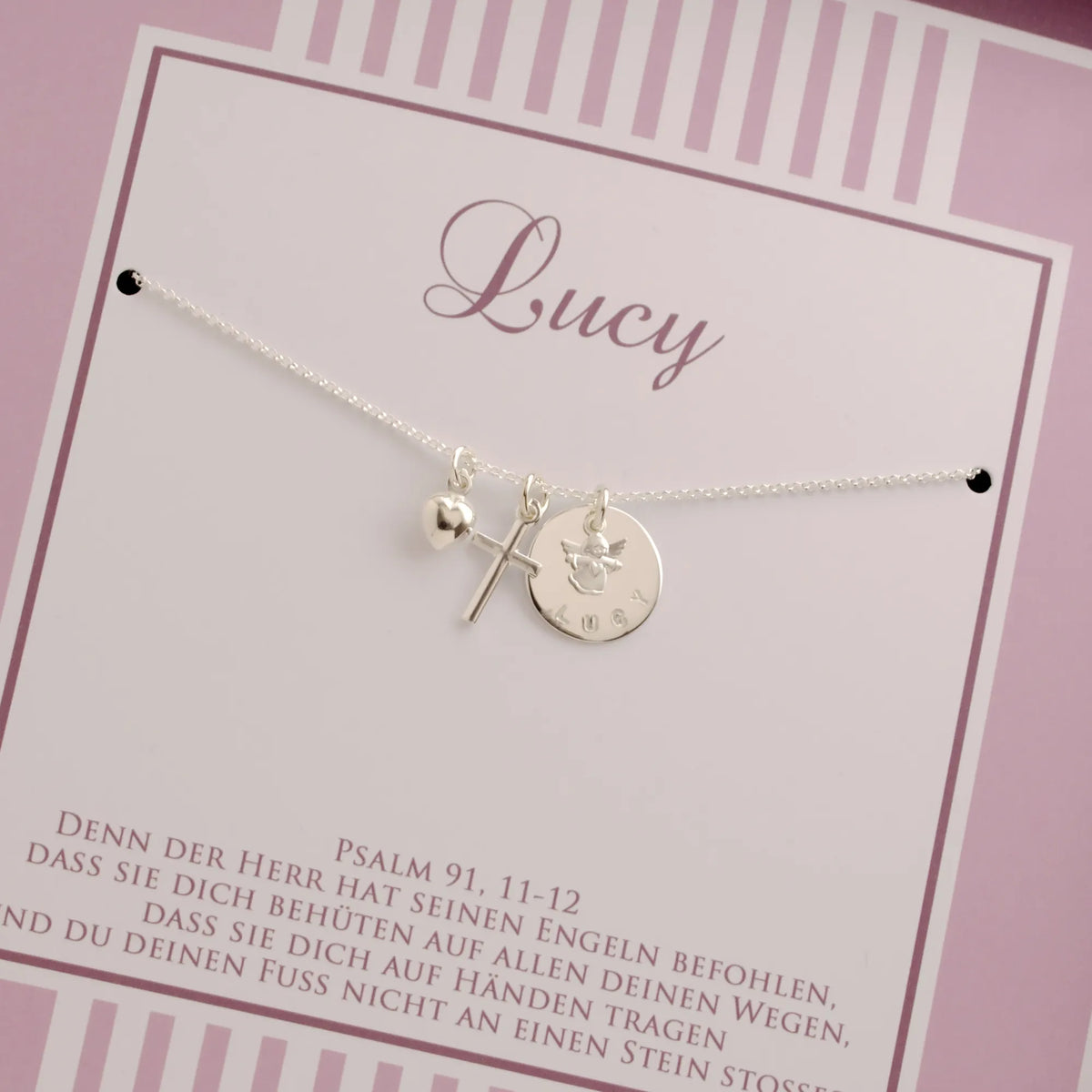 Detailansicht einer Karte mit feiner Silberkette mit Engel über einem Namensplättchen, Kreuz und Herz-Anhänger, personalisiert für 'Lucy'. Unterhalb der Kette ist der Taufspruch: Psalm 91,11-12 gedruckt.
