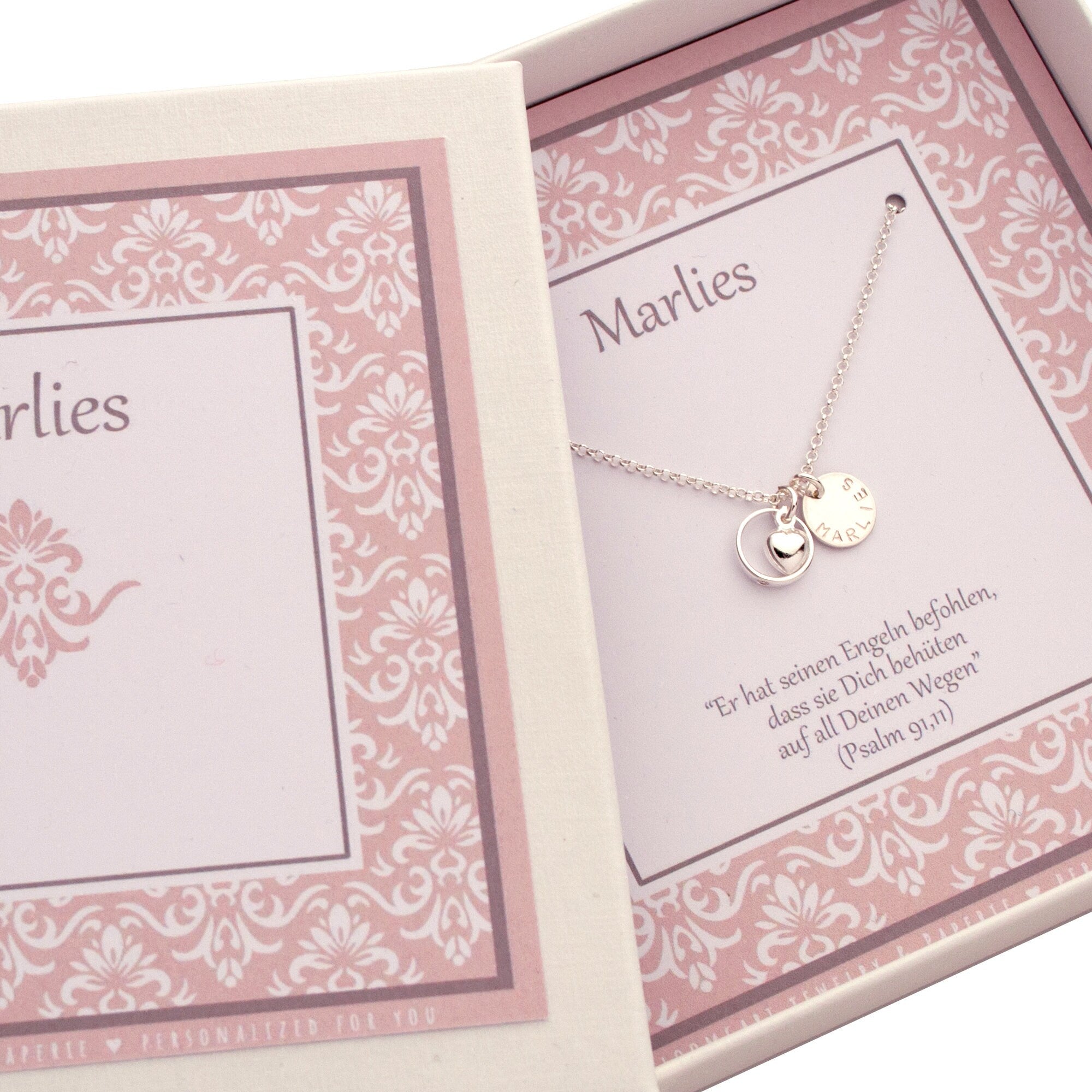 Die personalisierte Taufring-Kette "Stella" aus 925er Sterling Silber liegt liebevoll präsentiert in einer weißen Geschenkbox von Bloomgart mit rosa-weißem Muster. Die Kette mit Taufring, Herz-Anhänger und Gravur-Plättchen ist auf einer Karte mit dem Namen "Marlies" und dem Taufspruch "Er hat seinen Engeln befohlen, dass sie Dich behüten auf all Deinen Wegen" (Psalm 91,11) drapiert.