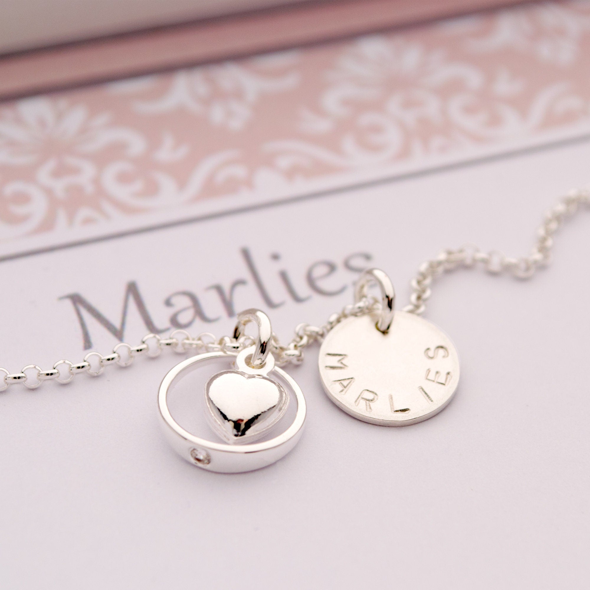 Nahaufnahme der personalisierten Taufring-Kette "Stella" aus 925er Sterling Silber. Die Kette zeigt einen Taufring mit einem kleinen Kristall und einem frei schwingenden Herz-Anhänger in der Mitte. Daneben hängt ein rundes Gravur-Plättchen mit dem Beispielnamen "Marlies". Handgefertigter Schmuck von Bloomgart als liebevolles Geschenk zur Taufe.