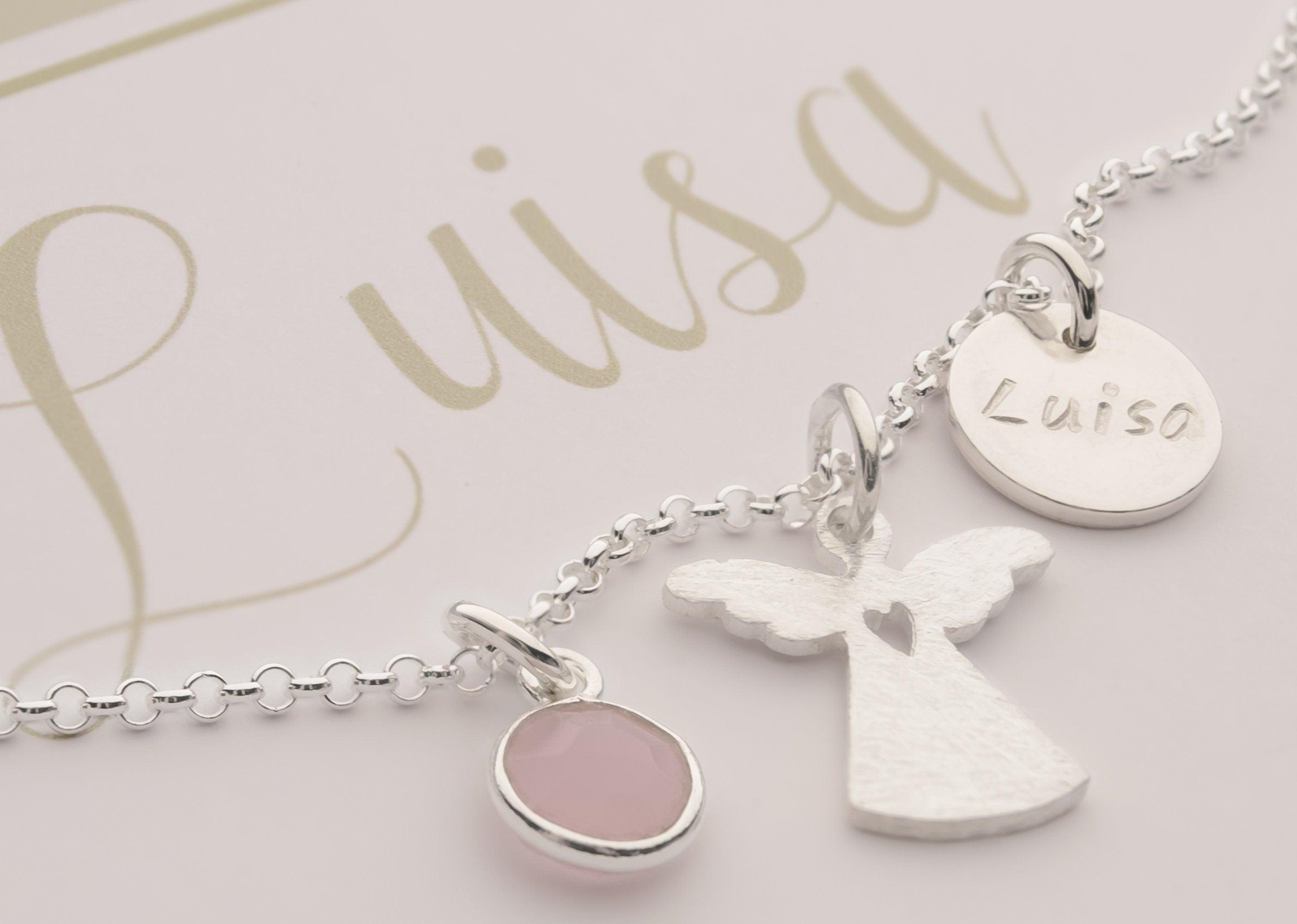 Nahaufnahme einer personalisierten Halskette aus 925 Silber. Die Kette ist mit einem filigranen Engel mit Herz Cutout, einem Rosenquarz und einem Plättchen mit der Namensgravur 'Luisa' versehen.