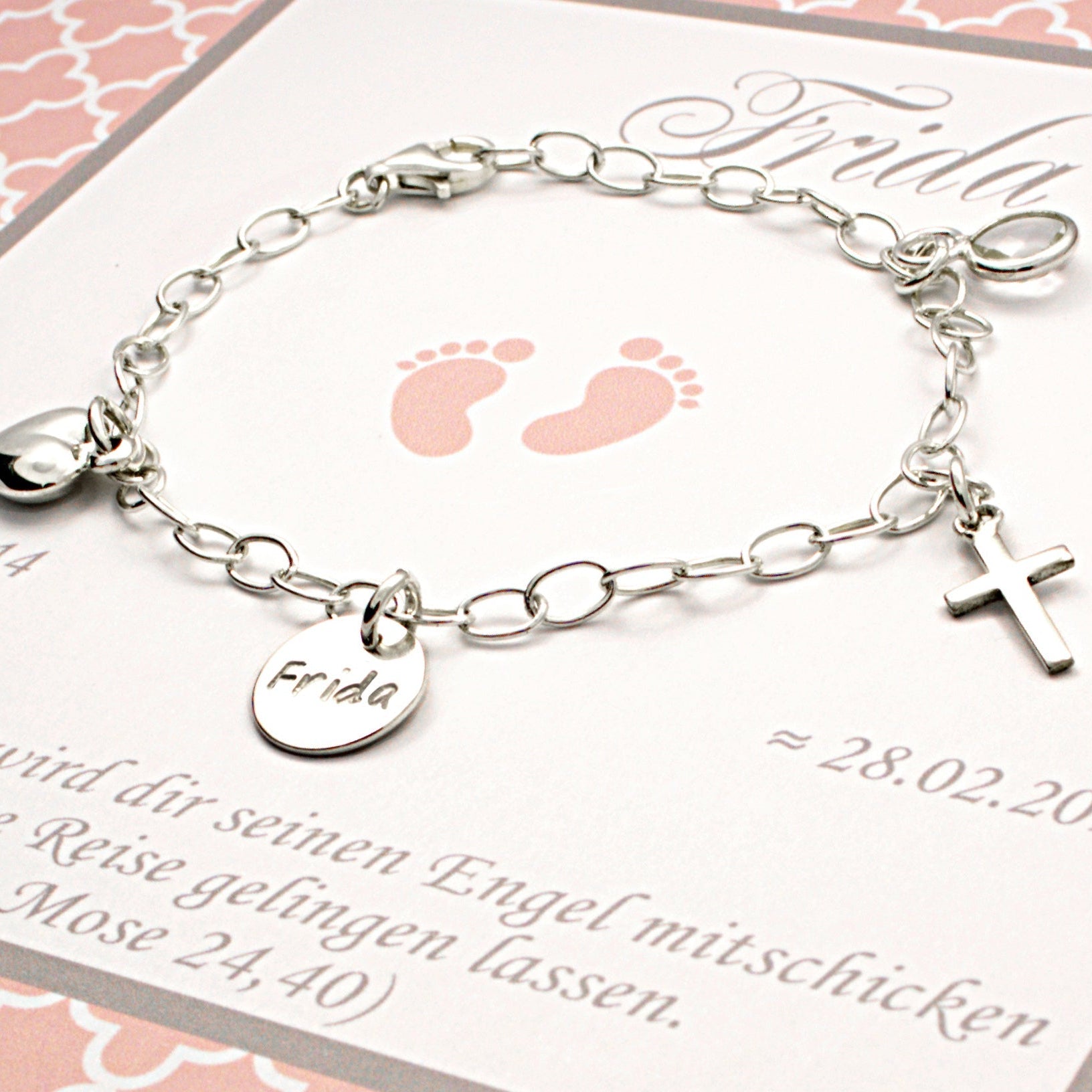 Detailansicht eines Silberarmbands mit vier Anhängern: Herz, Namentaler 'Frida', Kreuz und Geburtsstein auf rosa Karte.