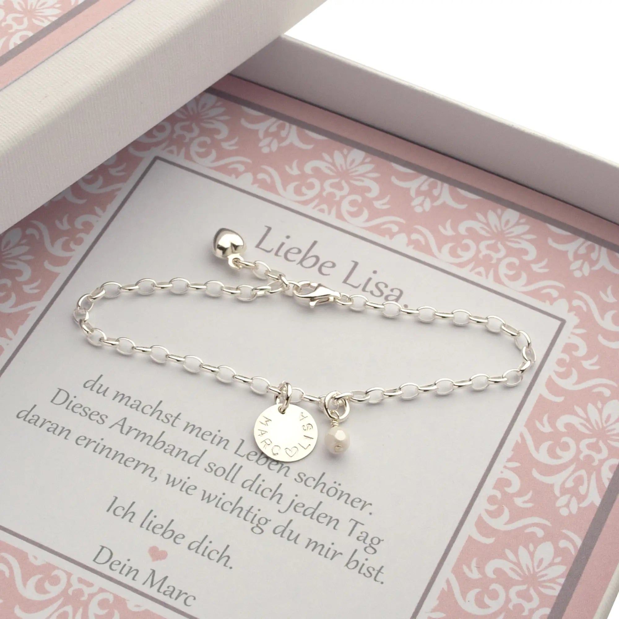 Eine personalisierte Geschenkbox mit weiß-rosa Ornamentmuster für Lisa.  Auf einer personalisierten Karte liegt ein 925 Silberarmband mit Namenstaler "Lisa ♡ Marc" und weißer Perle. Das Armband wird zum Geburtstag an die Ehefrau verschenkt.