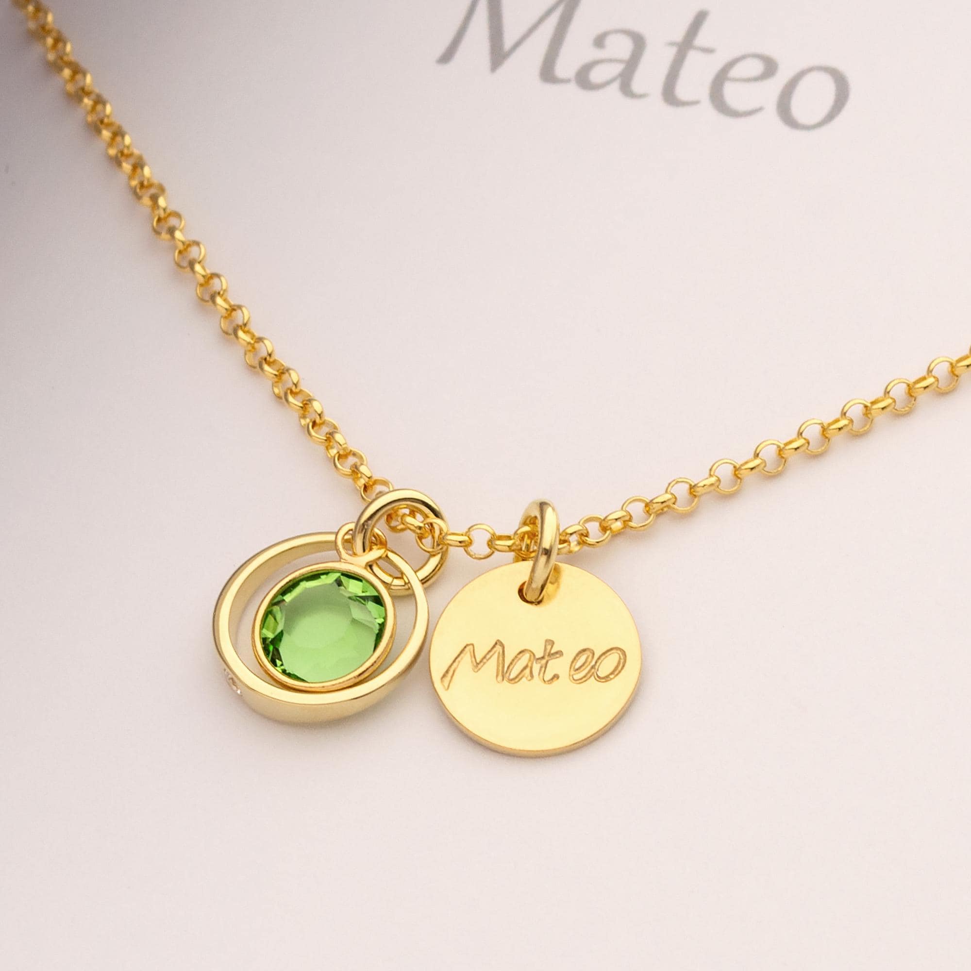 Nahaufnahme einer Gold Taufkette "Mateo" mit Namensgravur Taler und grünem Peridot-Geburtsstein in einem zierlichen Taufring mit weißem Zirkon.