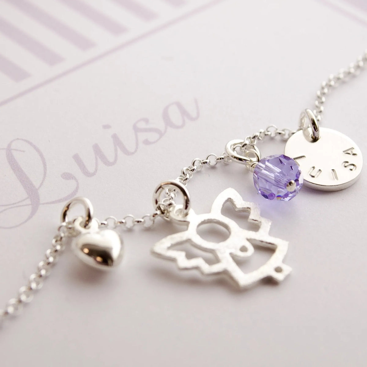 Detailansicht einer Taufkette mit Engel, Glücksstein in flieder, Herz und Namenstaler in personalisierter Geschenkbox mit Streifenmuster in weiß-flieder. Ein zauberhaftes Geschenk zur Taufe für Mädchen von ihren Paten