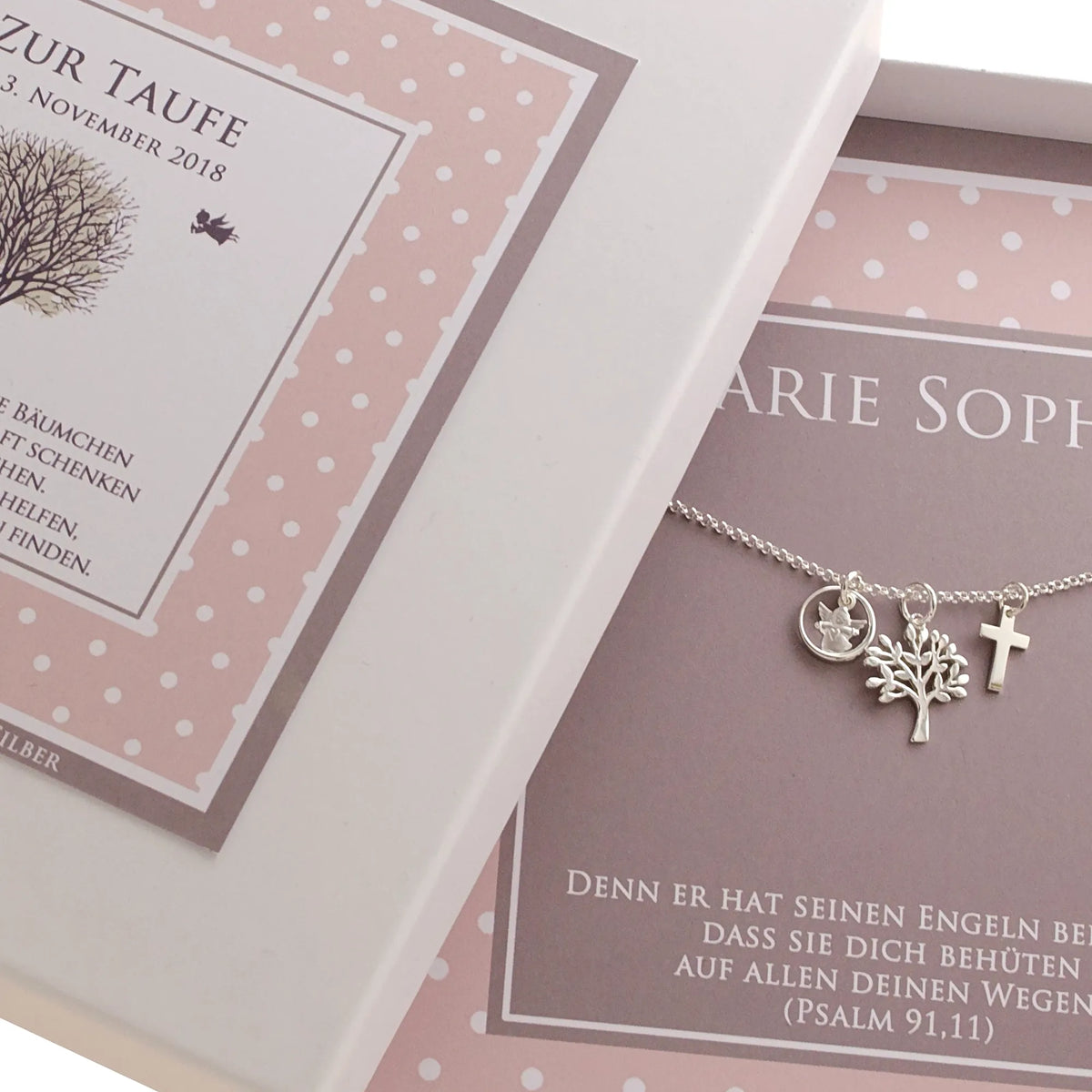 Angeschnittene Darstellung einer personalisierten Geschenkbox im rosa-taupe Pünktchen-Design zur Taufe. Innenliegend befindet sich eine zarte Gliederkette mit Engel im Ring, Lebensbaum und Kreuz.