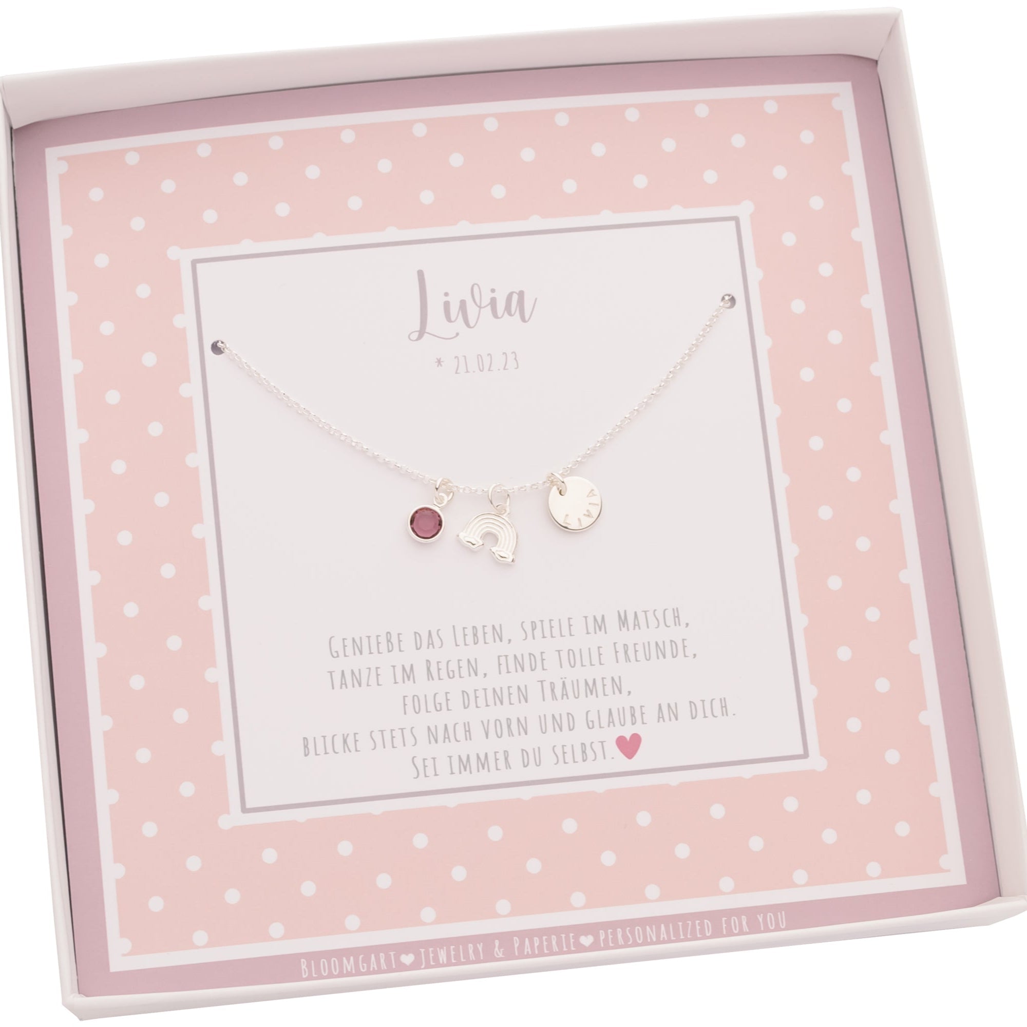 Innenansicht einer Geschenkbox mit rosa-weißem Punktemuster. Innen liegt eine filigrane Kette aus 925 Silber mit Namenstaler, Regenbogen und Geburtsstein.