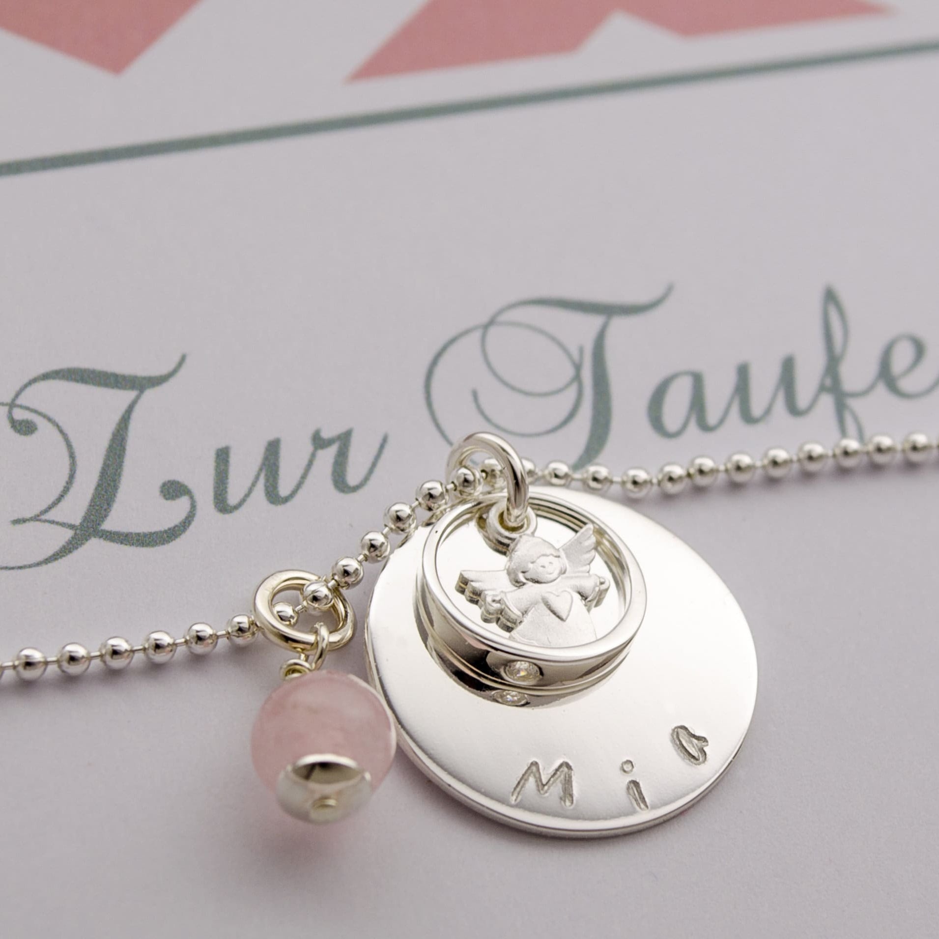 Nahaufnahme der Taufkette "Mia" aus 925 Silber, die den gravierten Namens-Taler, einen kleinen Schutzengel im Taufring und einen Rosenquarz-Anhänger zeigt.