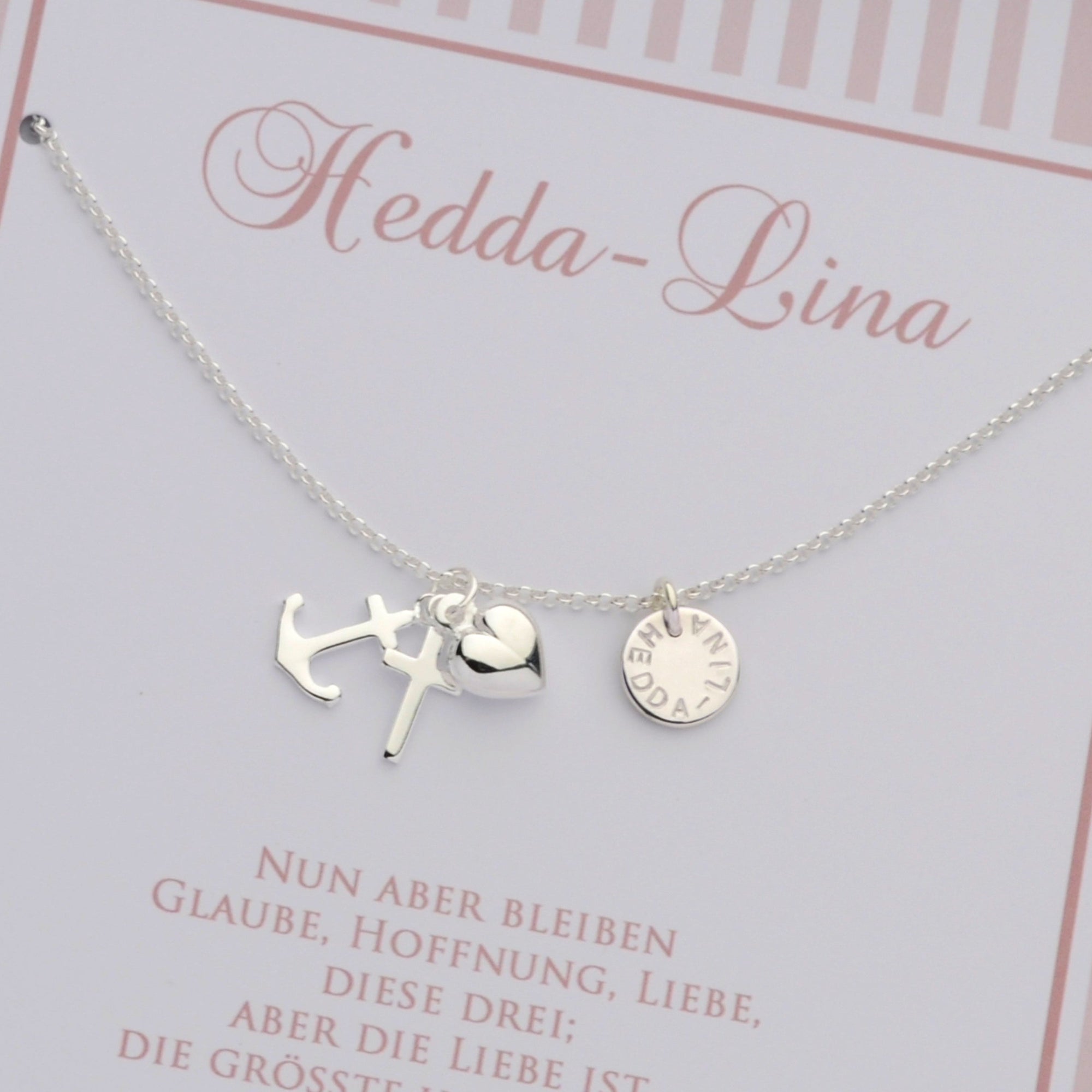 Detailansicht einer personalisierten Silberkette mit Anhängern für Glaube (Kreuz), Liebe (Herz) und Hoffnung (Anker) sowie einem Gravurtaler mit dem Namen "Hedda-Lina" auf einer rosa Karte montiert.
