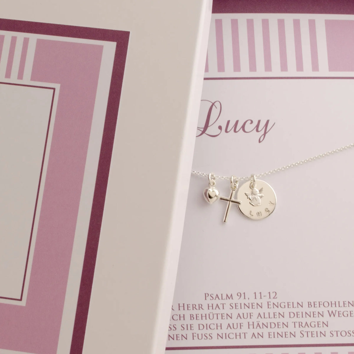 Detailansicht einer rosa Geschenkbox mit filigranem Taufschmuck aus 925 Silber mit Schutzengel, Kreuz und Herz-Anhänger, personalisiert für 'Lucy'. Unterhalb der Kette ist der Taufspruch: Psalm 91,11-12 gedruckt.