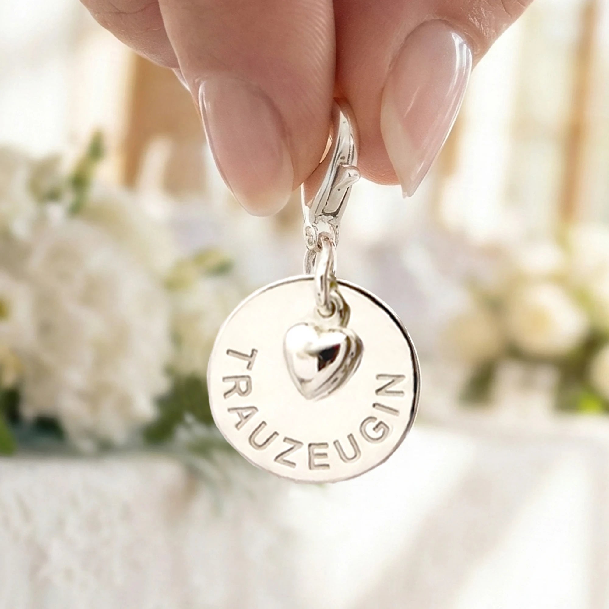 Nahansicht eines Trauzeugin-Gravur-Anhängers mit Karabinerhaken und Herz aus 925 Sterling Silber. Der Anhänger wird von einer Frau vor einem festlich geschmücktem Tisch präsentiert.