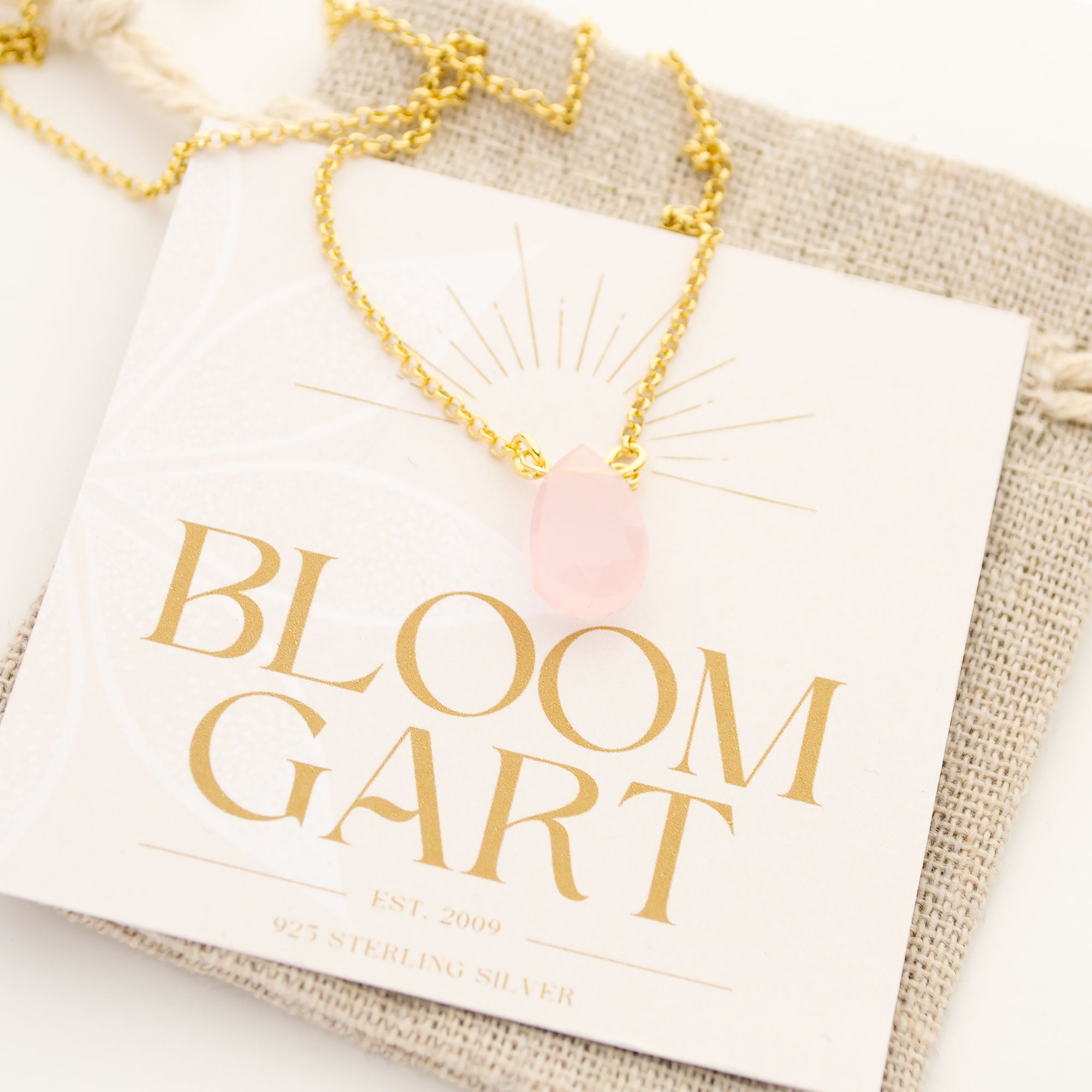 Nahaufnahme einer filigranen Goldkette mit einem hellrosa Rosenquarz Tropfen-Anhänger, der mit Draht an der Kette befestigt ist.