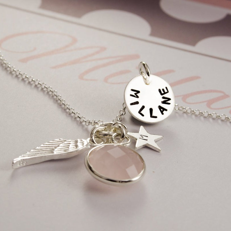 Engelsrufer Kette aus 925 Silber mit Flügel, Rosenquarz, Initial-Stern und Namensplättchen. Im Hintergrund sieht man einen Ausschnitt der handgemachten geschenkbox in rosa-weiß-beige, hier personalisiert für 'Millane'.